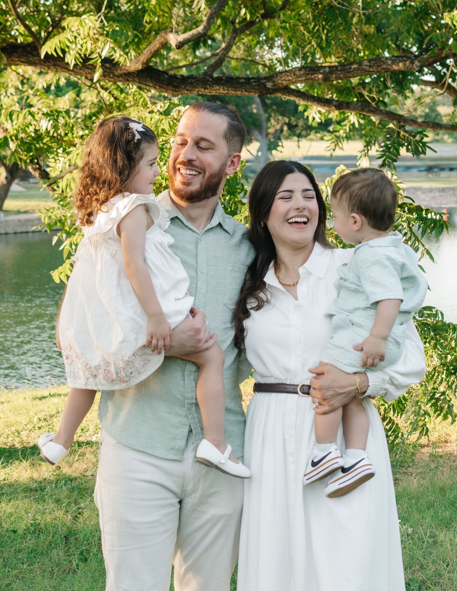 Happy family of four posing outdoors near a pond, parents holding two young children, dressed in light summer clothing.