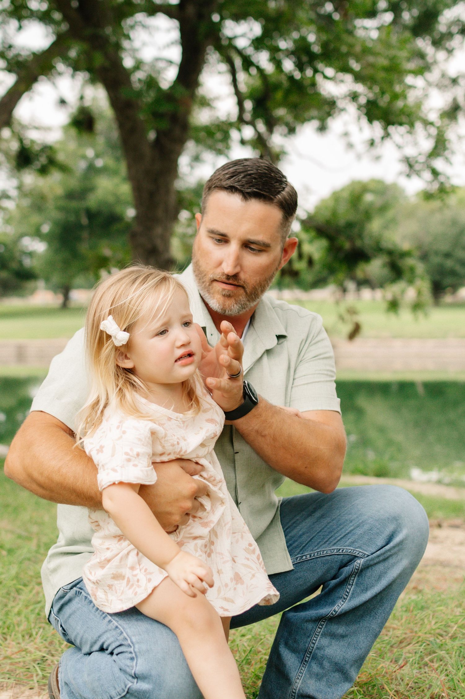 A father crouches outdoors holding his young blonde daughter in a park setting with green trees.