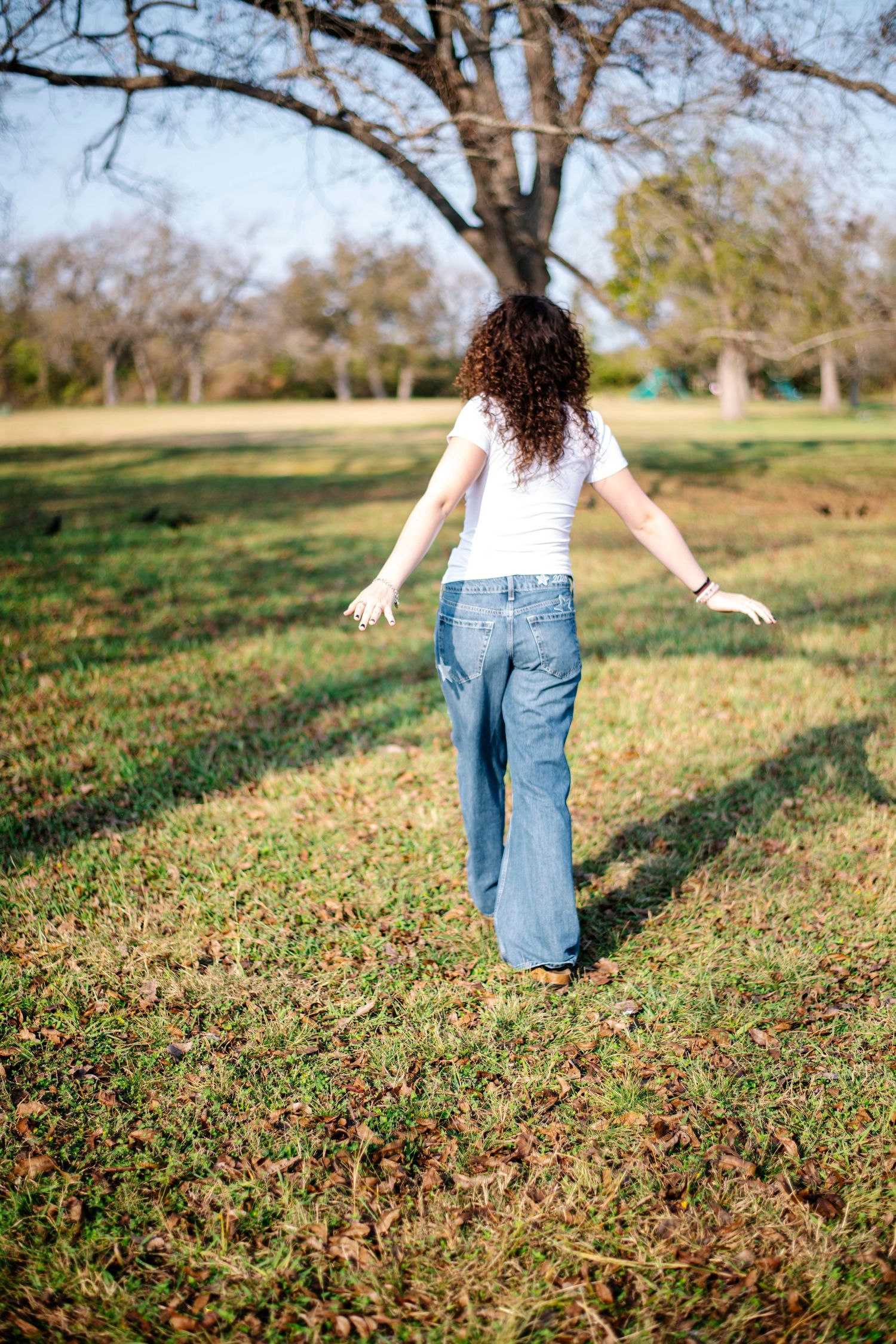 Young woman with curly hair in white top and jeans walks freely through a sunny green park with arms outstretched.