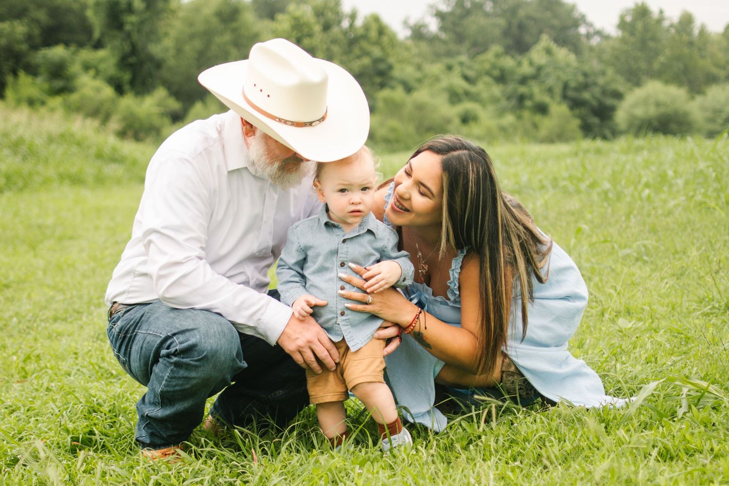 Family photo of grandparents and baby sitting in green grass field, man in cowboy hat kissing infant outdoors.