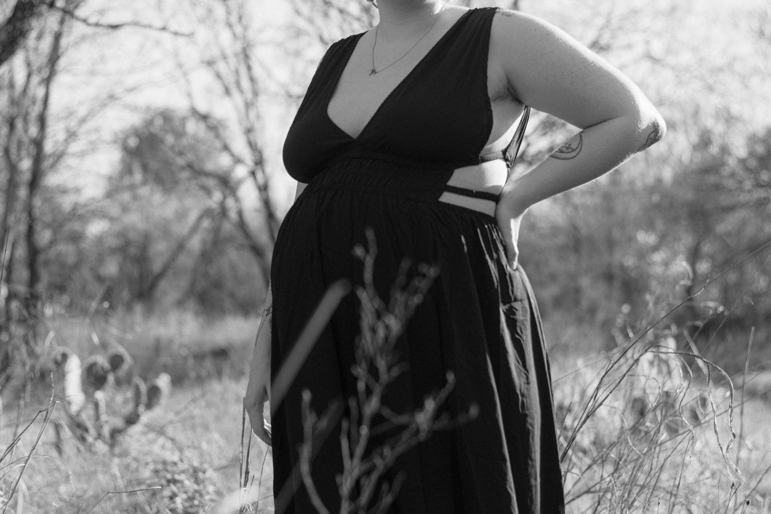 Black and white photo of a pregnant woman in a dark maxi dress posing outdoors among trees.