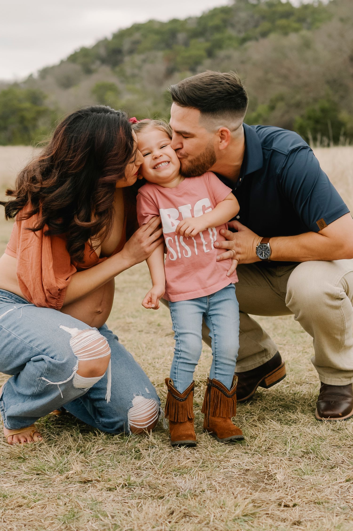 Parents kissing their toddler girl wearing a Big Sister shirt in a field, fun outdoor family photo.
