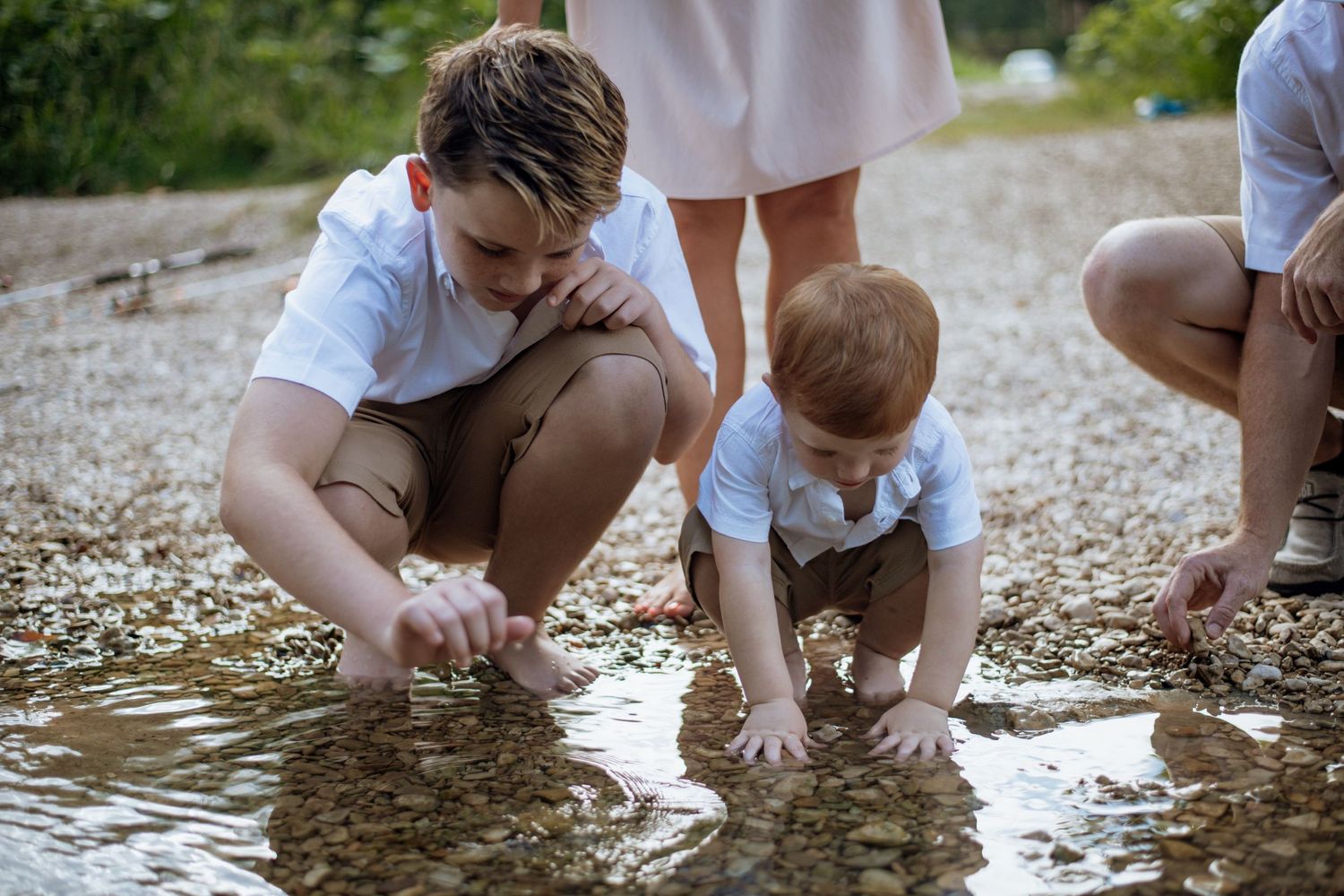 Two people in light-colored clothing crouch at the water's edge examining or playing with something in the shallow water.