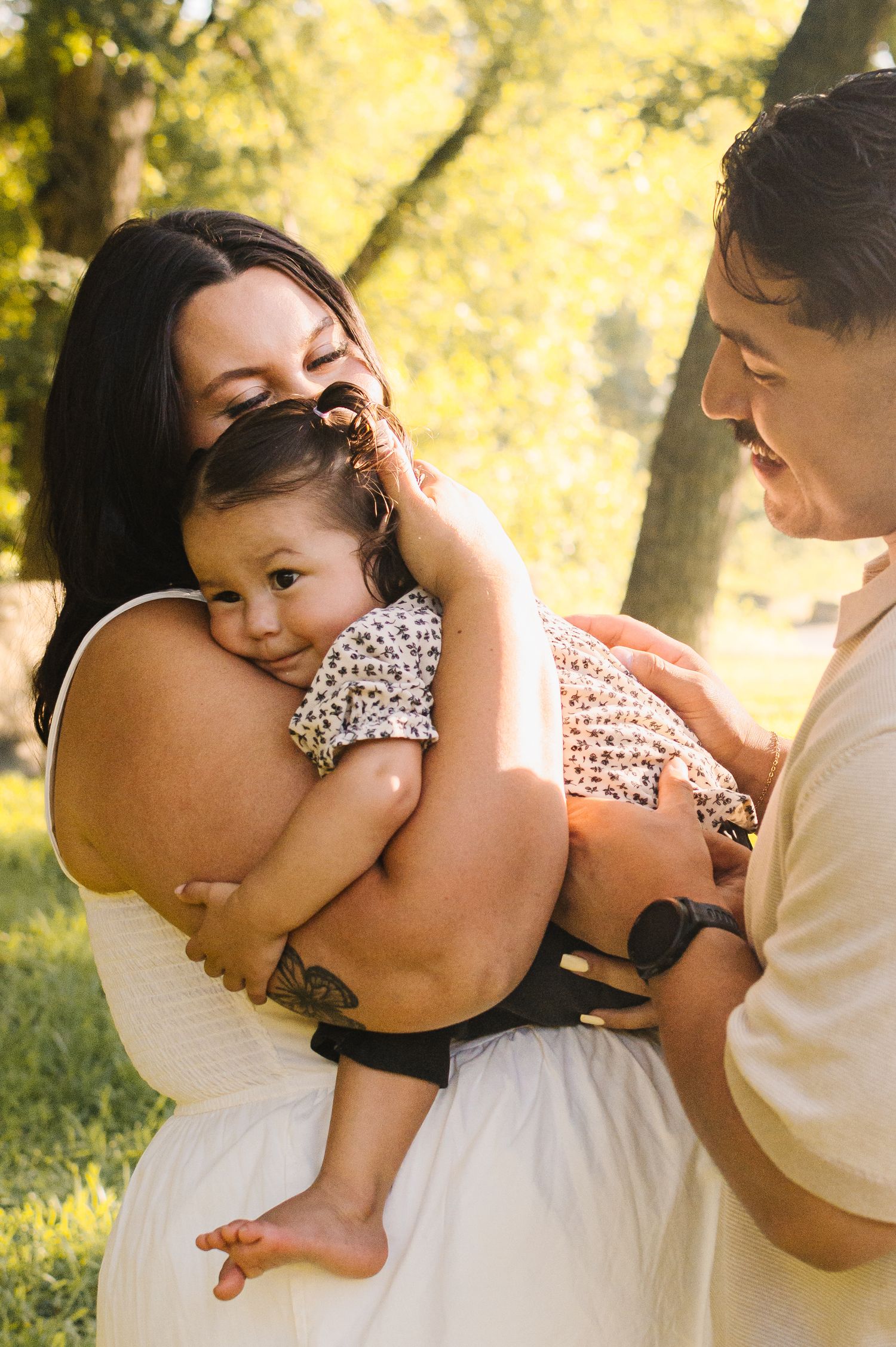 A young family shares a tender moment outdoors, with a mother in white holding a baby while a man smiles beside them.
