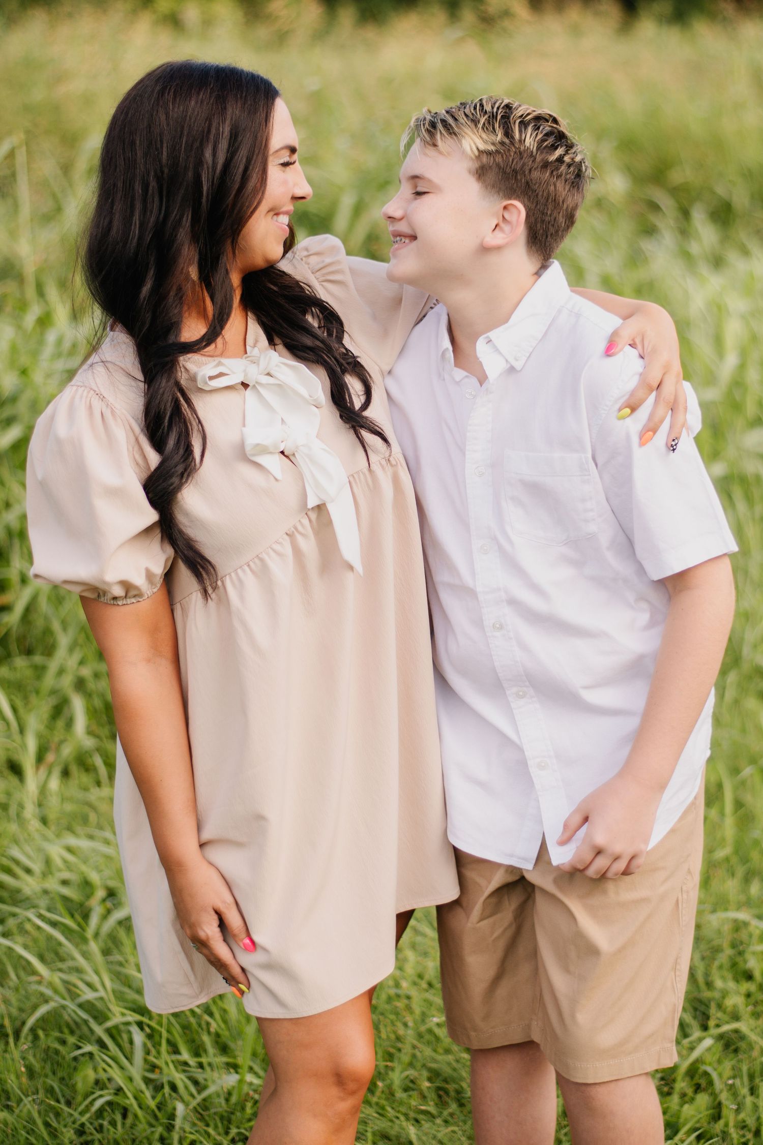 A woman in a beige dress and a young boy in a white shirt smile at each other in a grassy field.