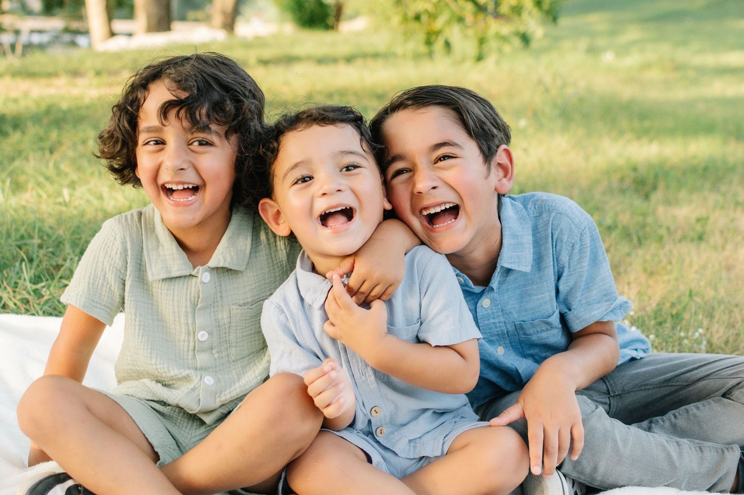 Three laughing boys sitting together on a blanket in a sunny green park, sharing a joyful moment outdoors.