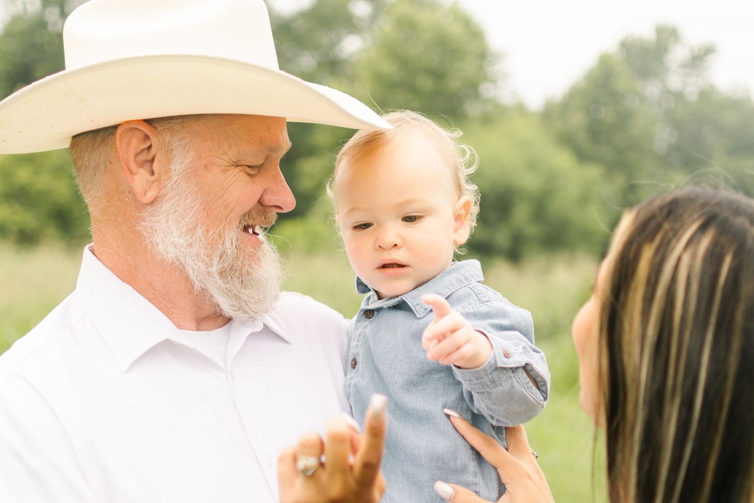An older man in a white cowboy hat holds a baby in a blue outfit outdoors in a green field.