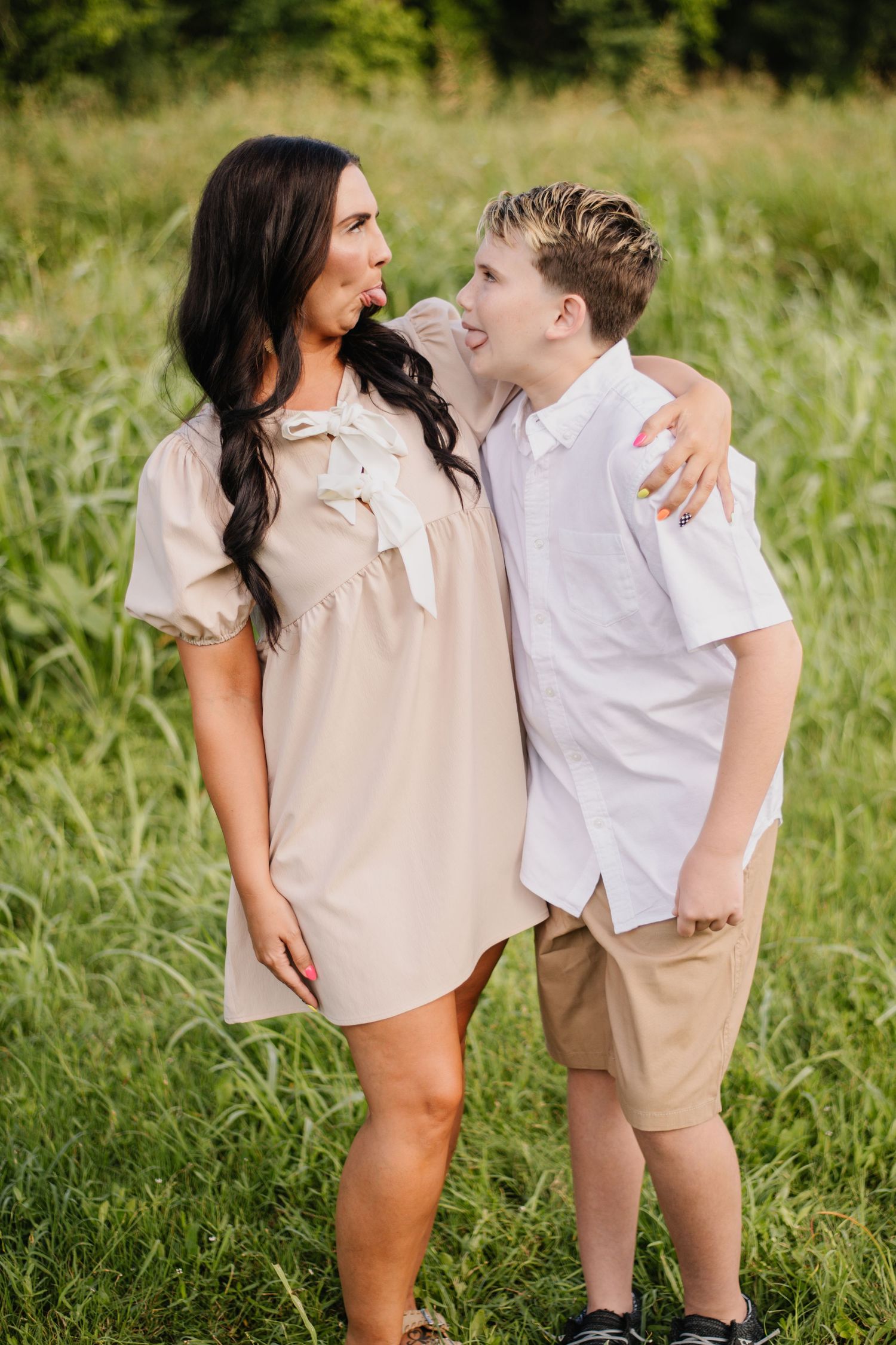 A woman in a beige dress and a boy in a white shirt share a playful moment outdoors in a green field.