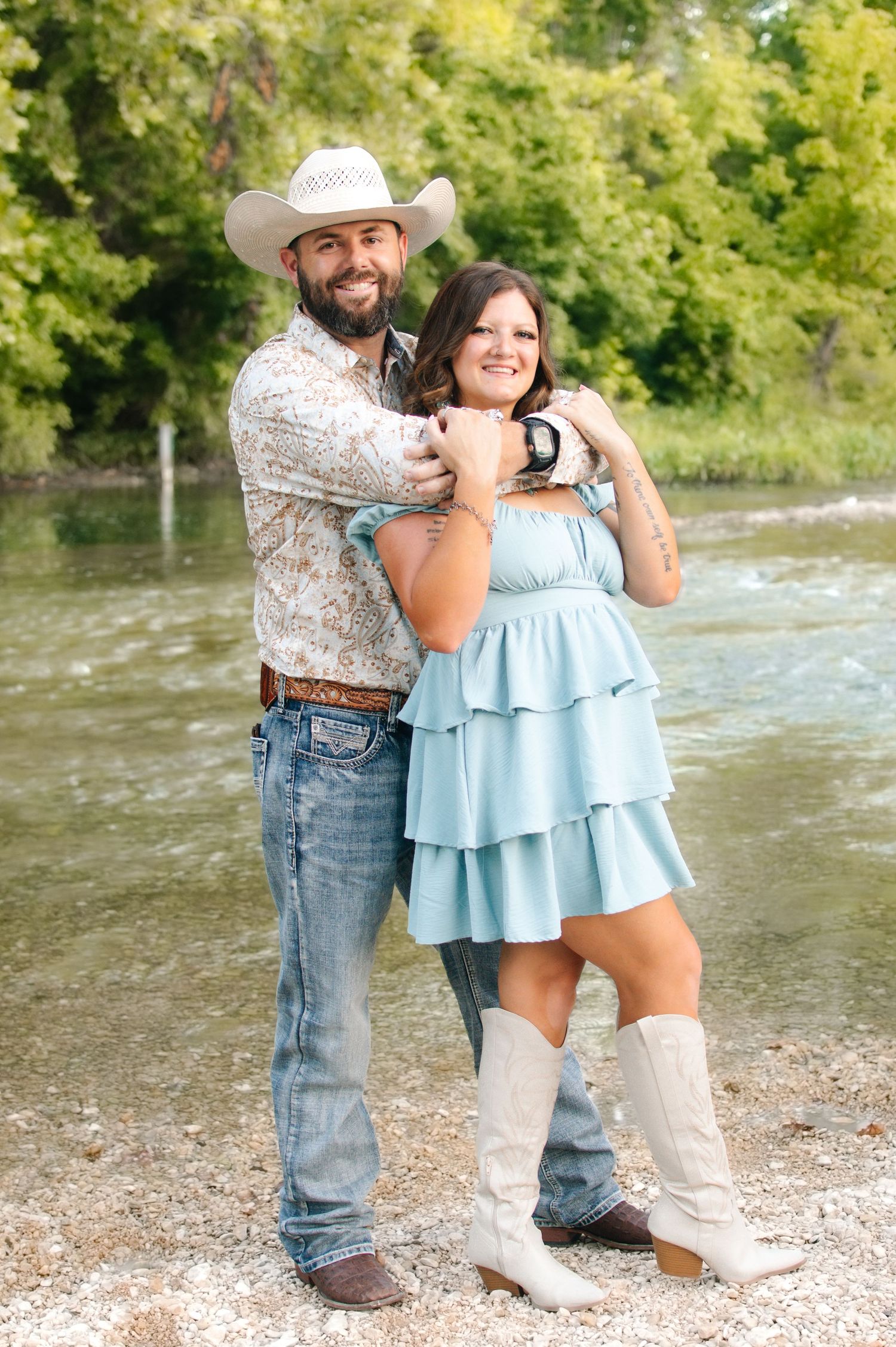 A couple poses by a river, man in cowboy hat and floral shirt, woman in blue ruffled dress and white cowboy boots.