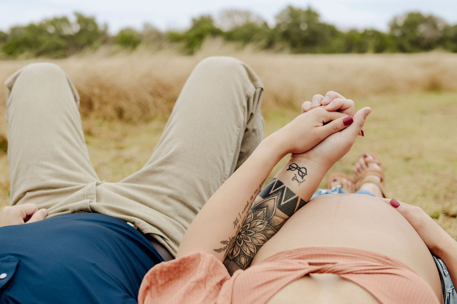 Pregnant couple lying in a field holding hands, woman with floral tattoo on arm wearing a peach top.
