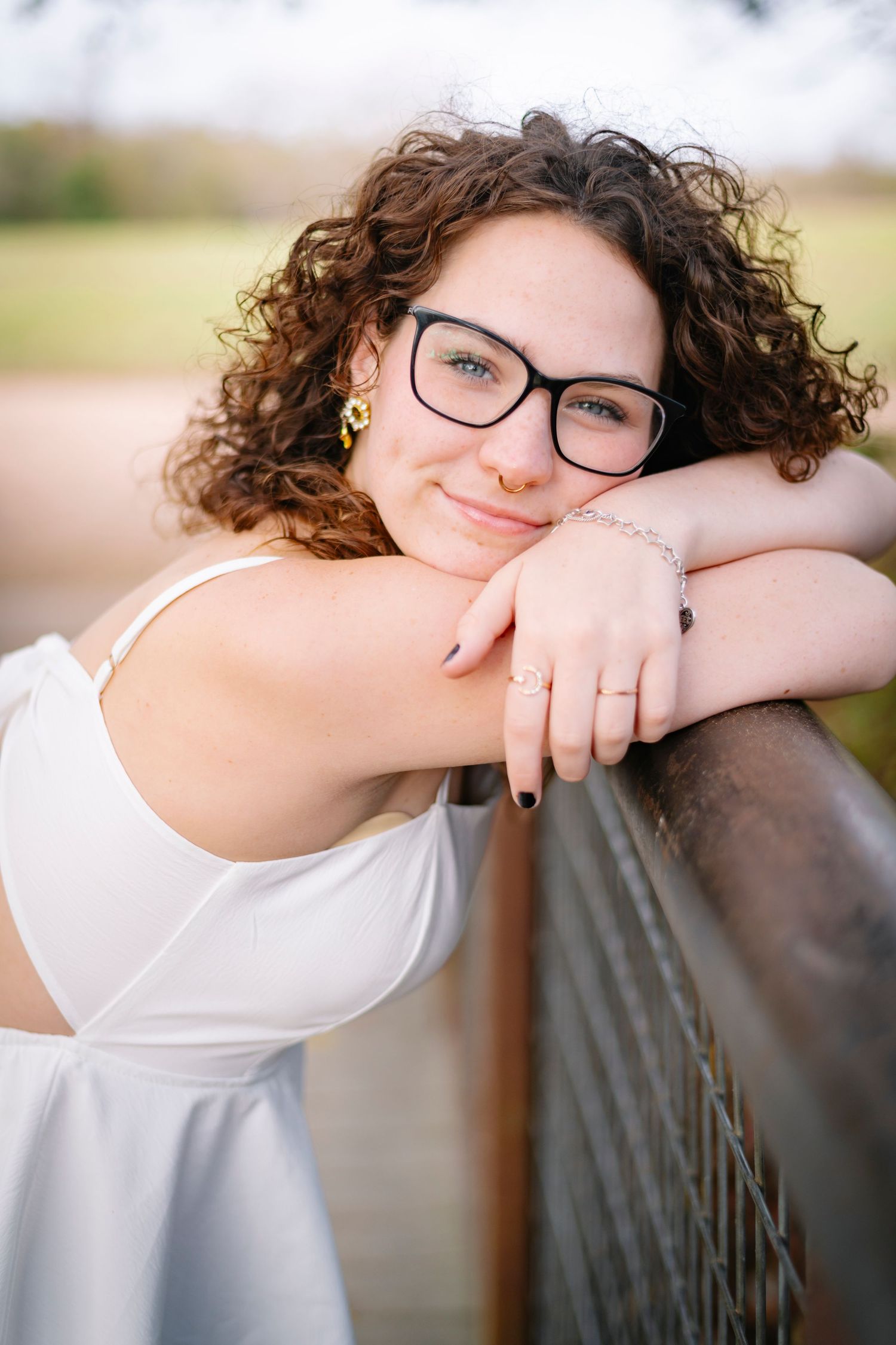Young woman with curly hair and glasses smiling while leaning on a wooden railing outdoors.