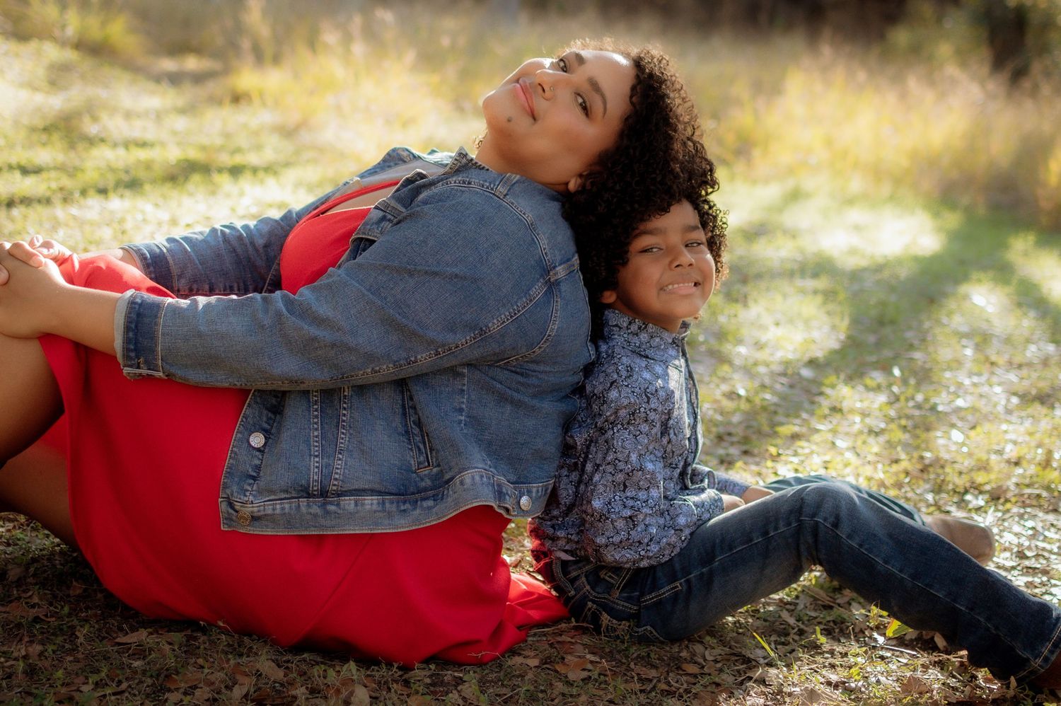 A mother and child sit back-to-back smiling outdoors in a sunny park, wearing casual denim and colorful clothing.