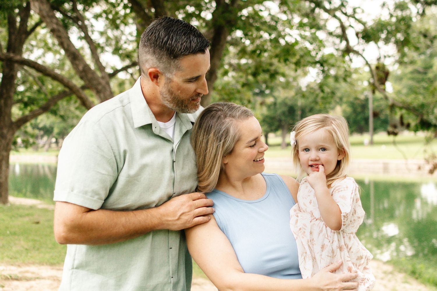 A happy family of three poses outdoors near a pond, with parents smiling at their young daughter.