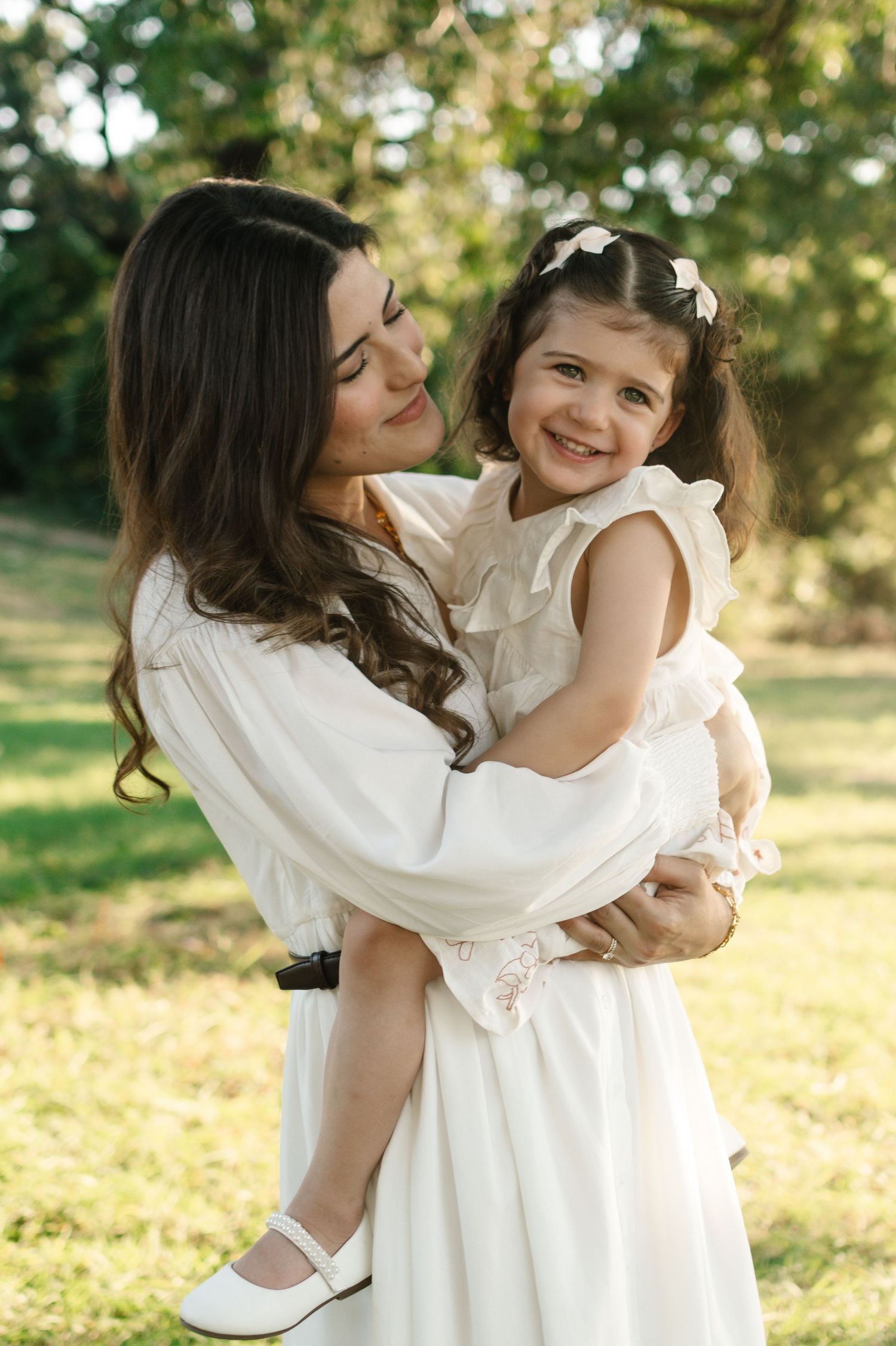 A mother in a white dress holds her smiling young daughter in a sunny green park, both wearing white outfits.