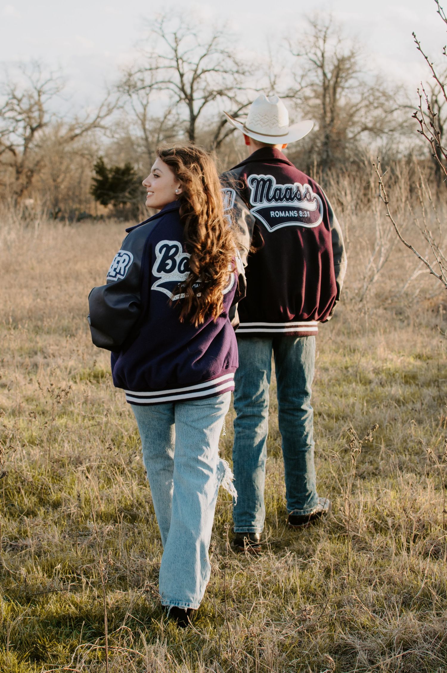 People wearing varsity jackets stand together in a grassy field during sunset.