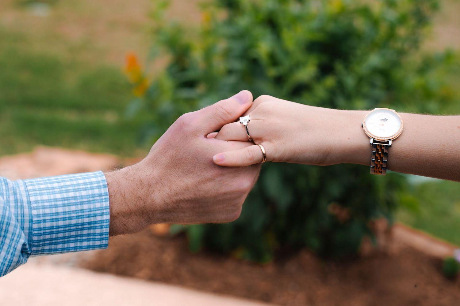 Close-up of couple holding hands outdoors, woman wearing elegant gold watch and diamond engagement ring.