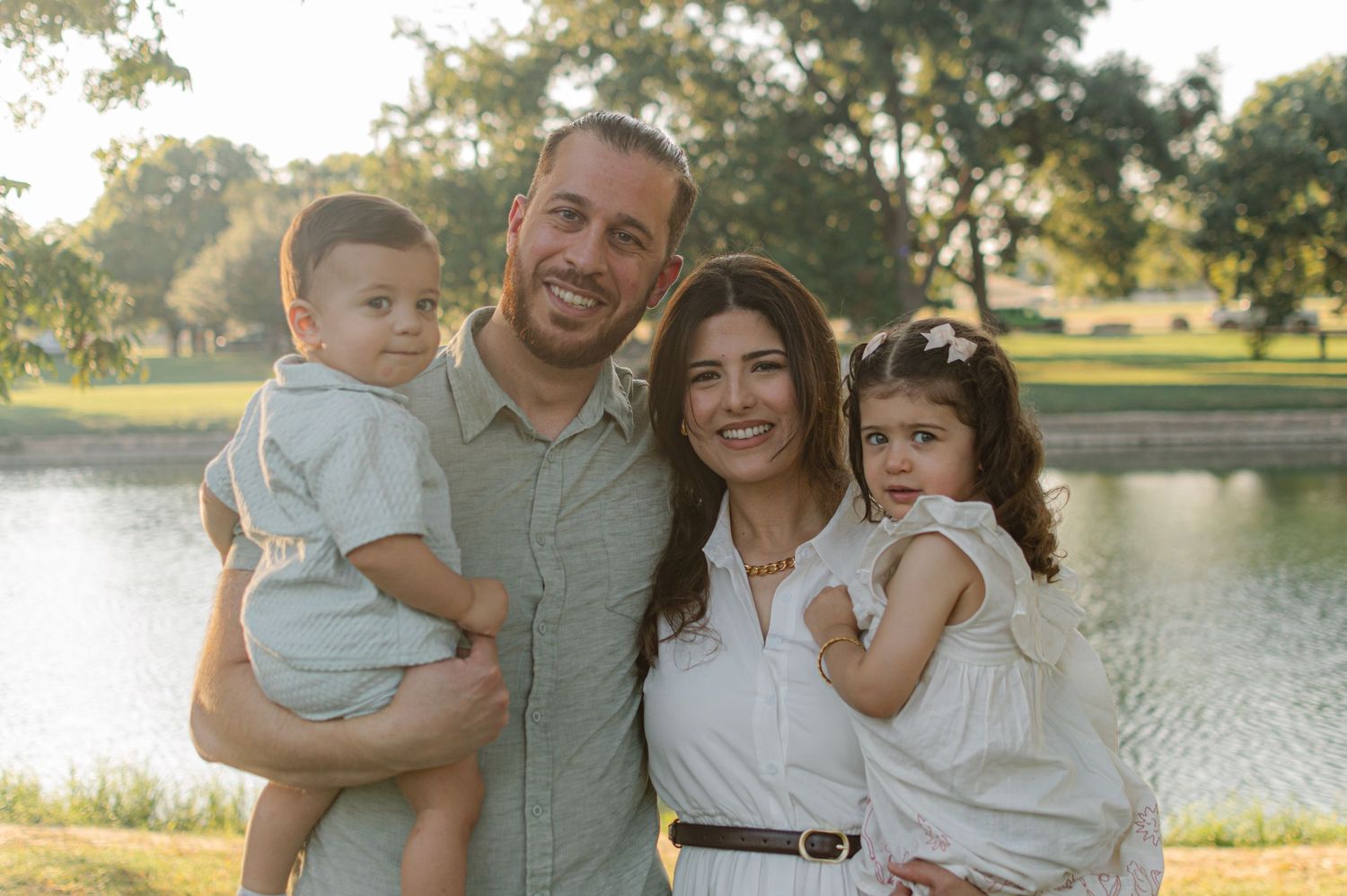 Happy family of four posing outdoors near a pond, parents holding young children, dressed in light colors on a sunny day.