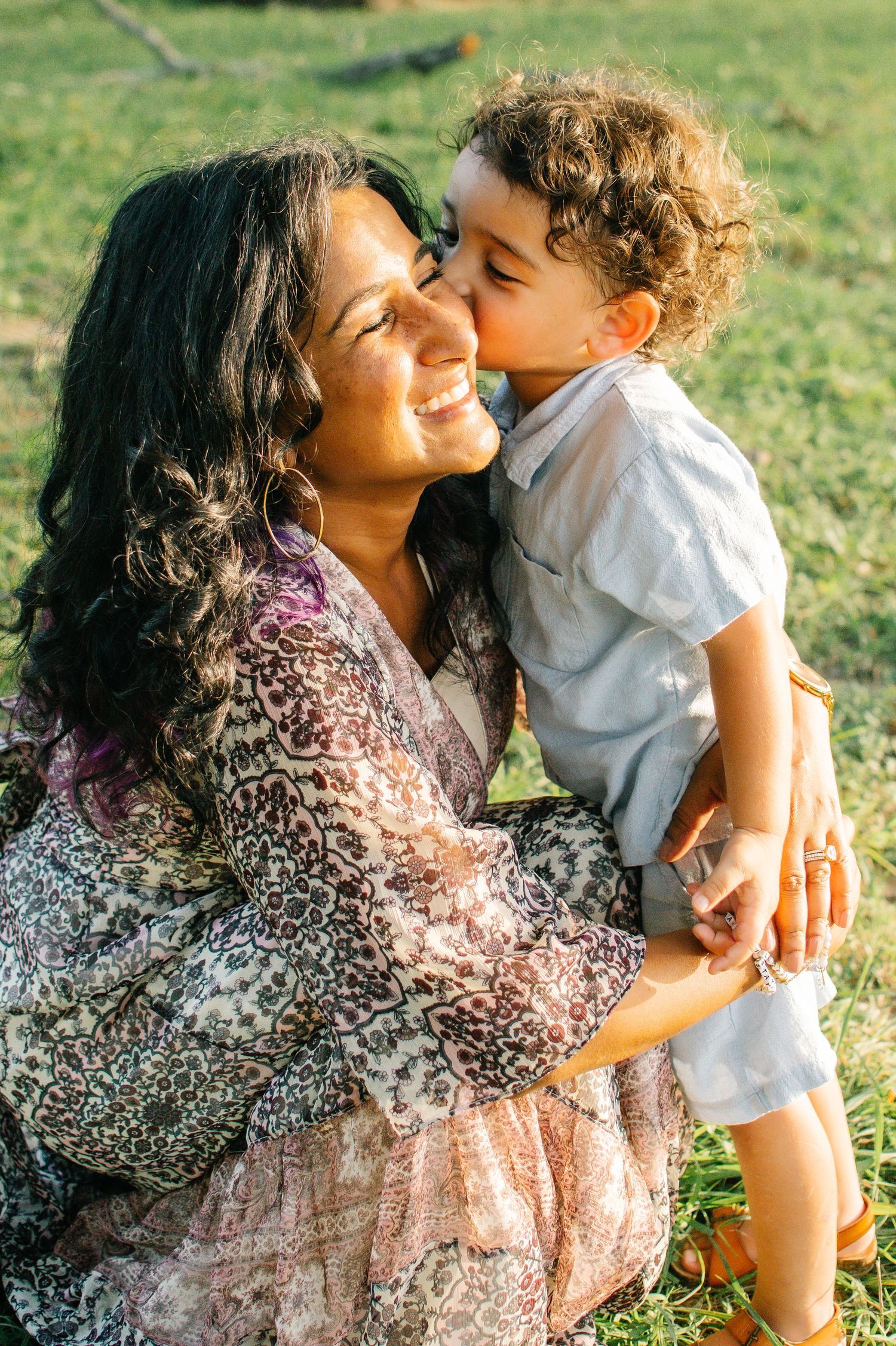 A smiling woman in a floral dress holds a curly-haired toddler boy who kisses her cheek outdoors.