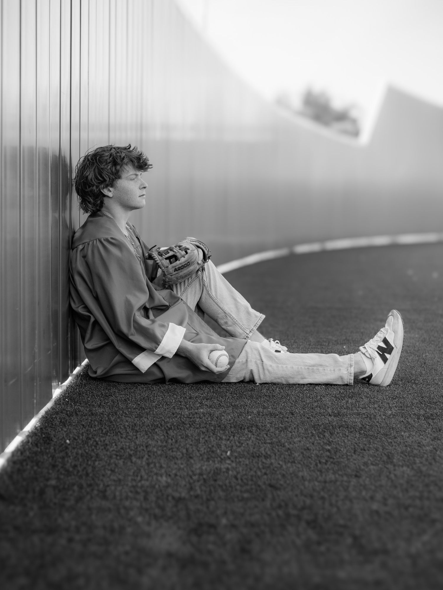 Black and white photo of a young man sitting against a metal wall on turf, wearing New Balance sneakers and holding a baseball glove.
