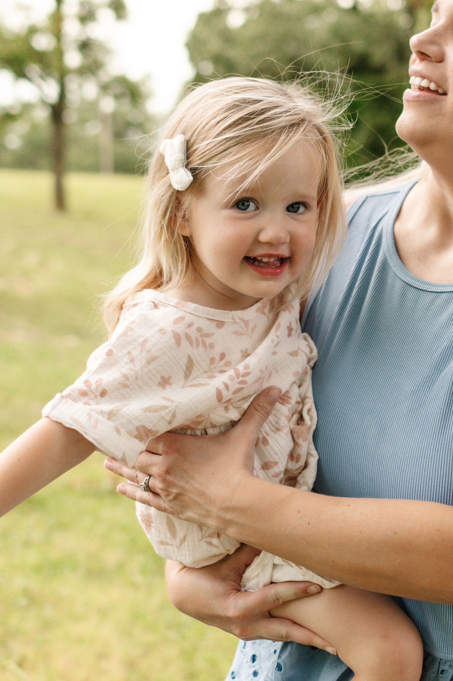 A smiling toddler girl with blonde hair and a white bow clip being held by a woman in a blue top outdoors.