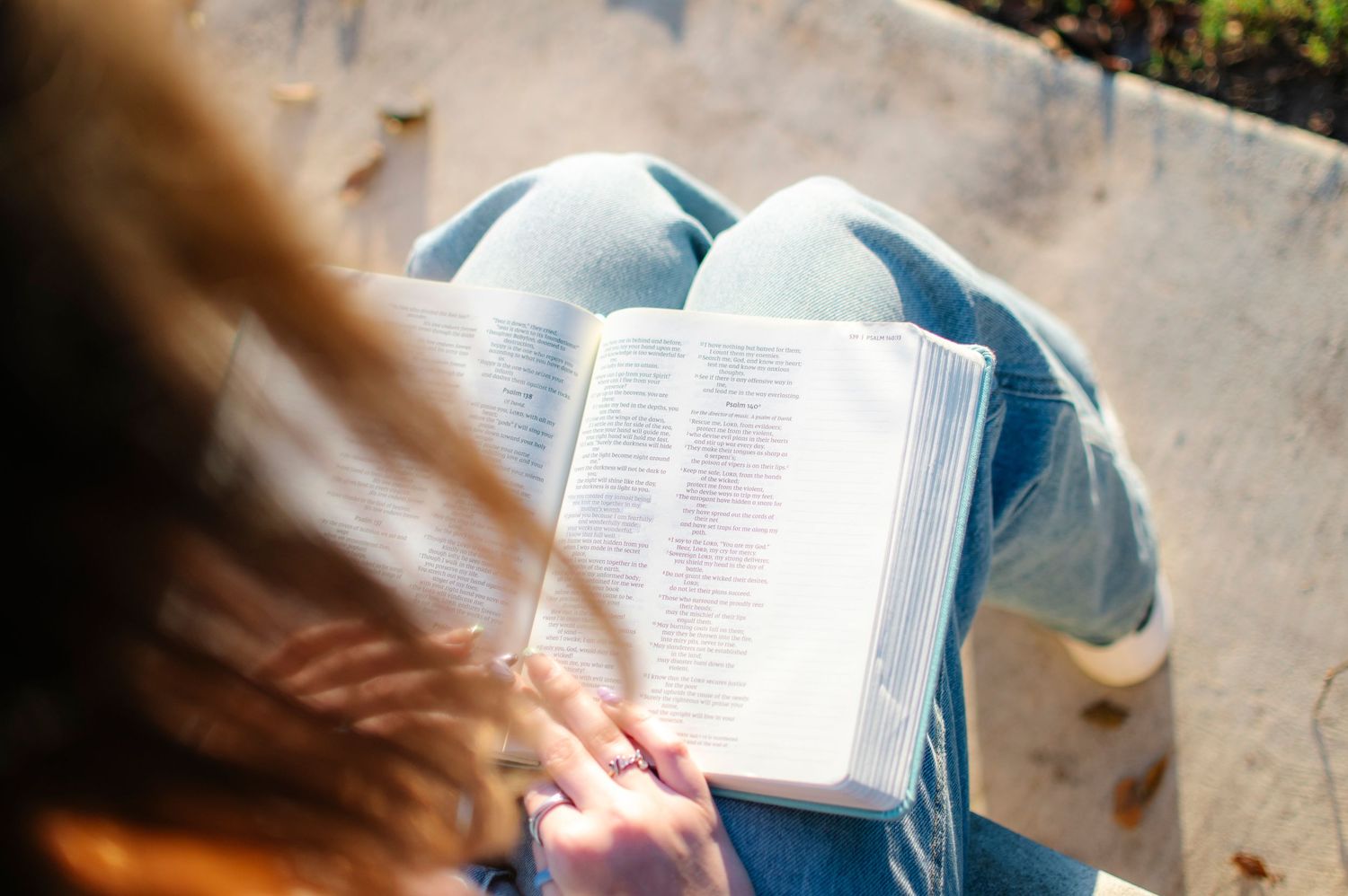 Young woman with long red hair reading an open book outdoors, seated on a bench in sunlight.