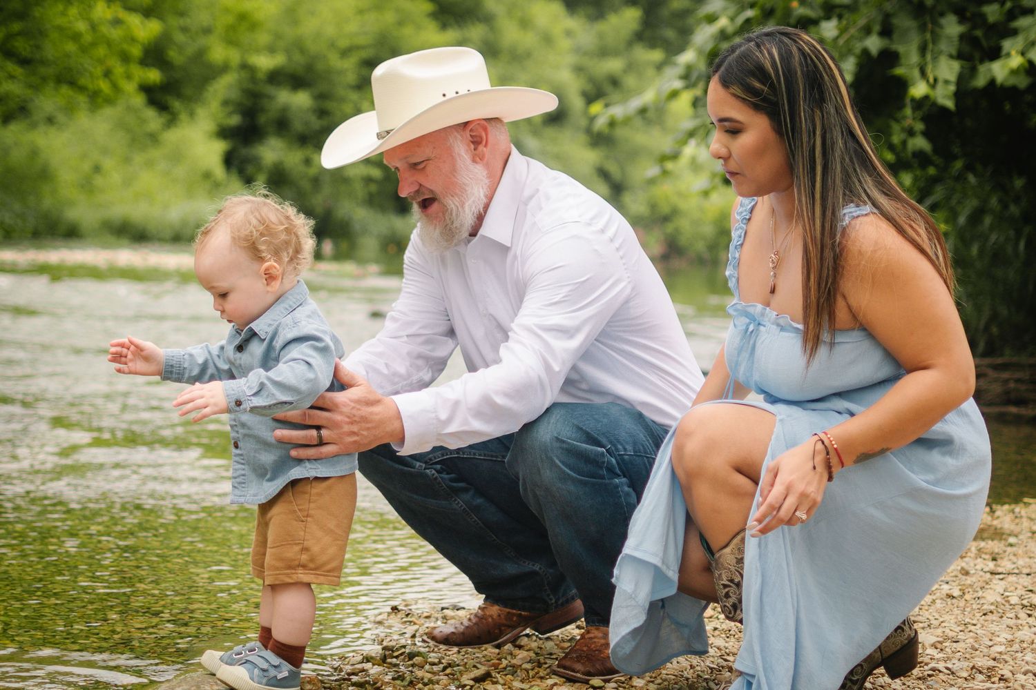 Family photo of grandpa in cowboy hat helping toddler walk by a creek while young woman watches.