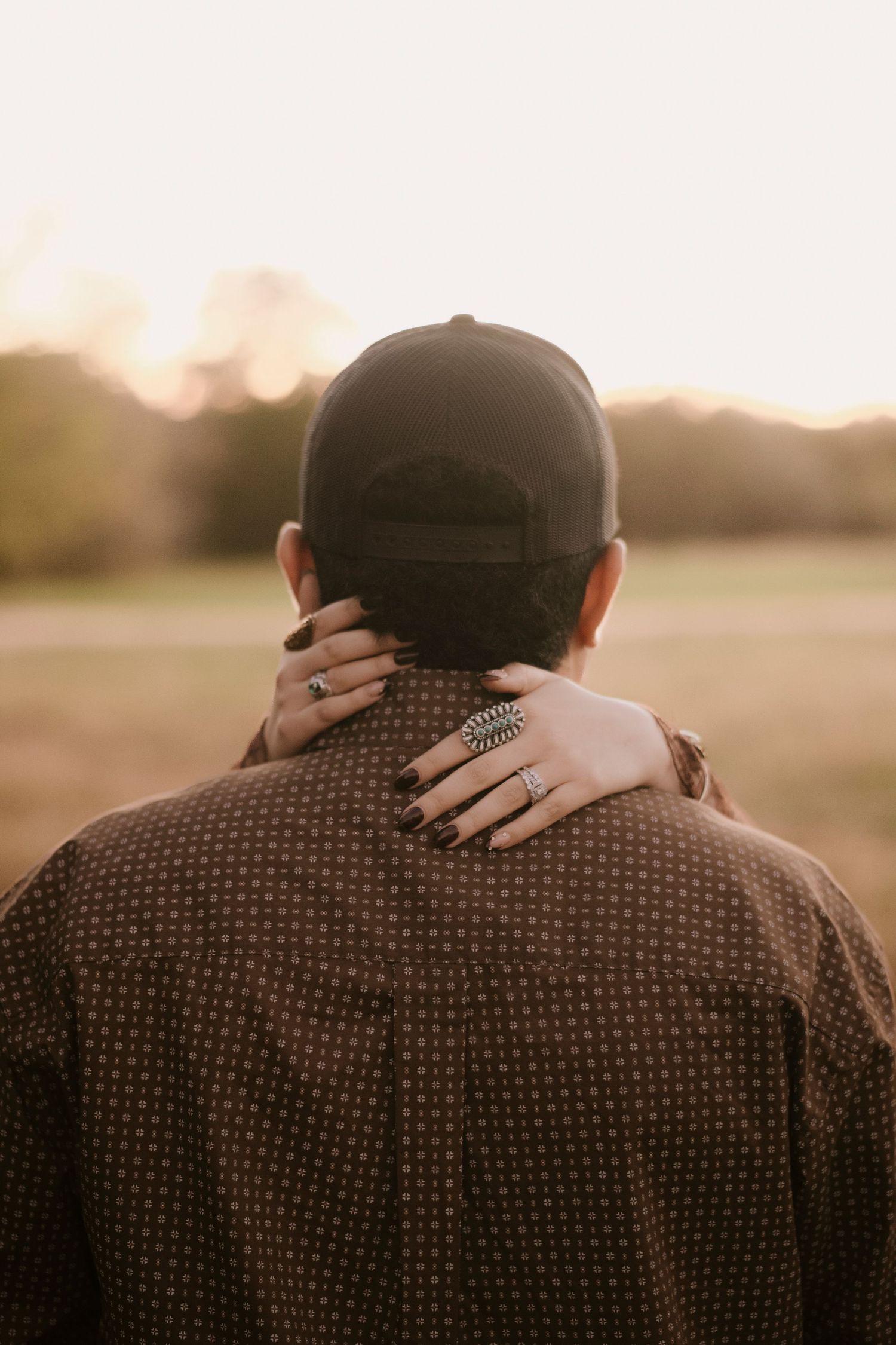 Person in brown polka dot shirt and black cap embraced from behind by hands with dark nails and silver rings at sunset.