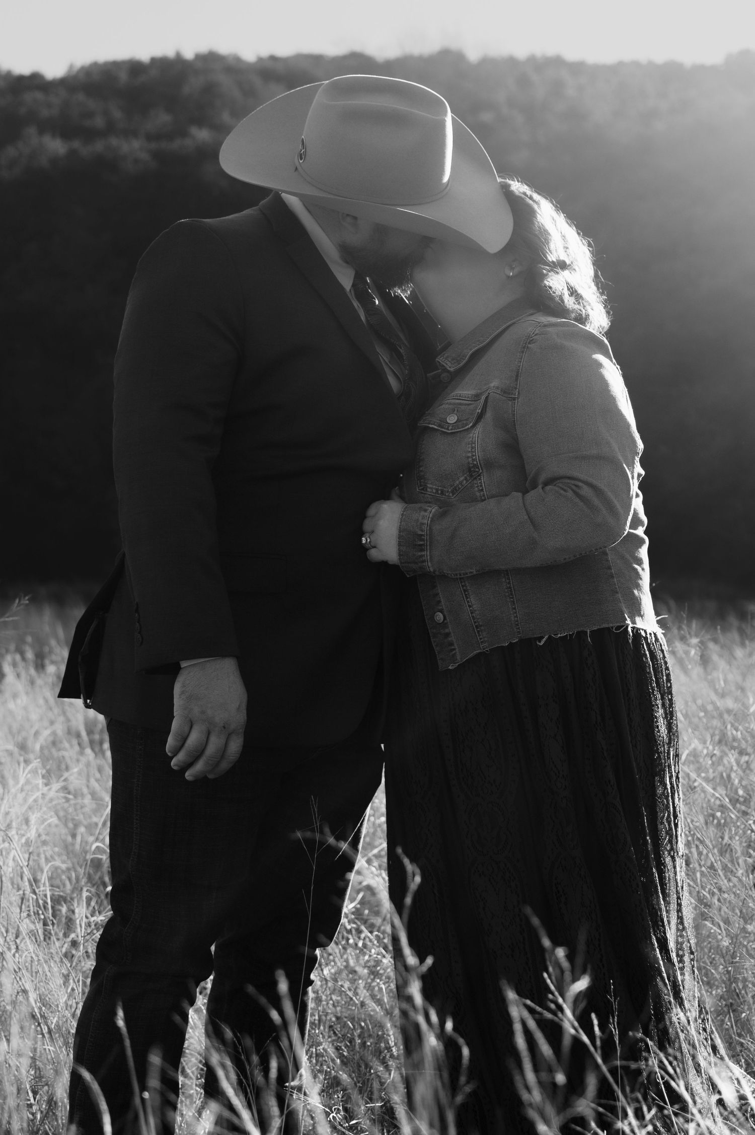 Black and white photo of a couple kissing in a field, man wearing cowboy hat and suit, woman in denim jacket and skirt.