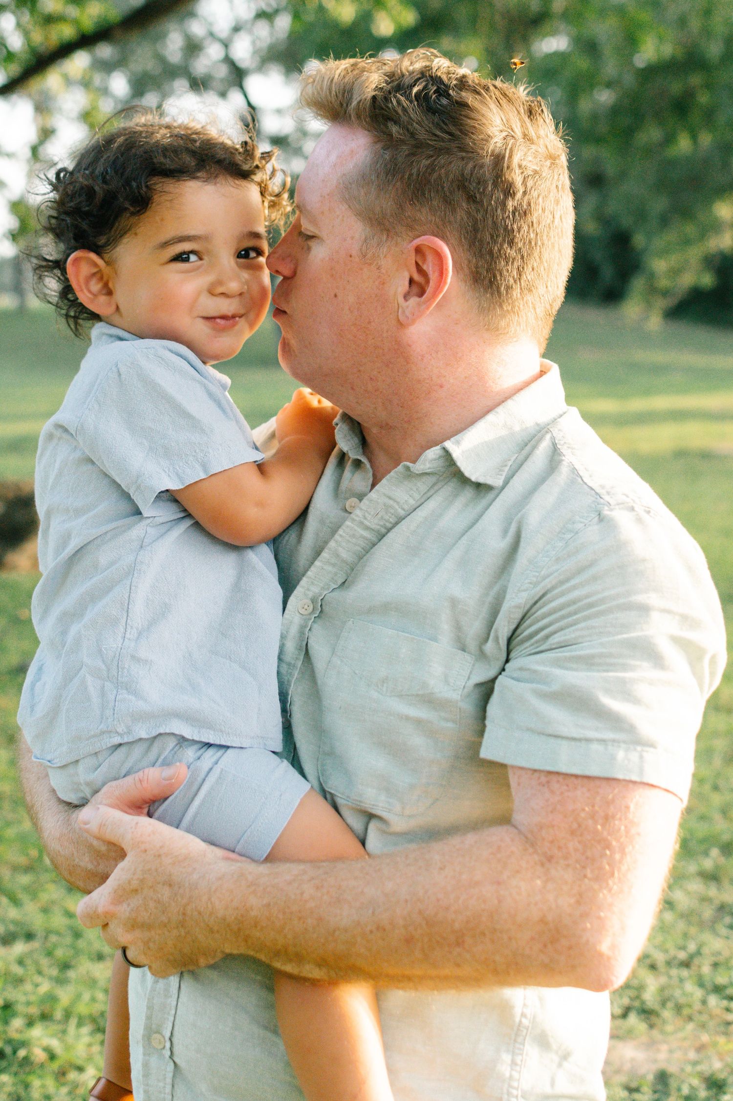 A loving father kisses his toddler son on the cheek in a sunlit park.