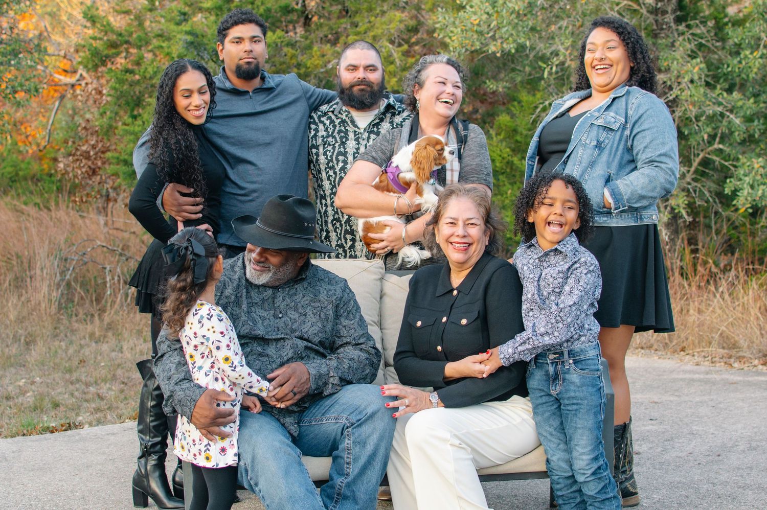 Multi-generational family portrait outdoors in autumn, eight members smiling together, one holding a small dog.