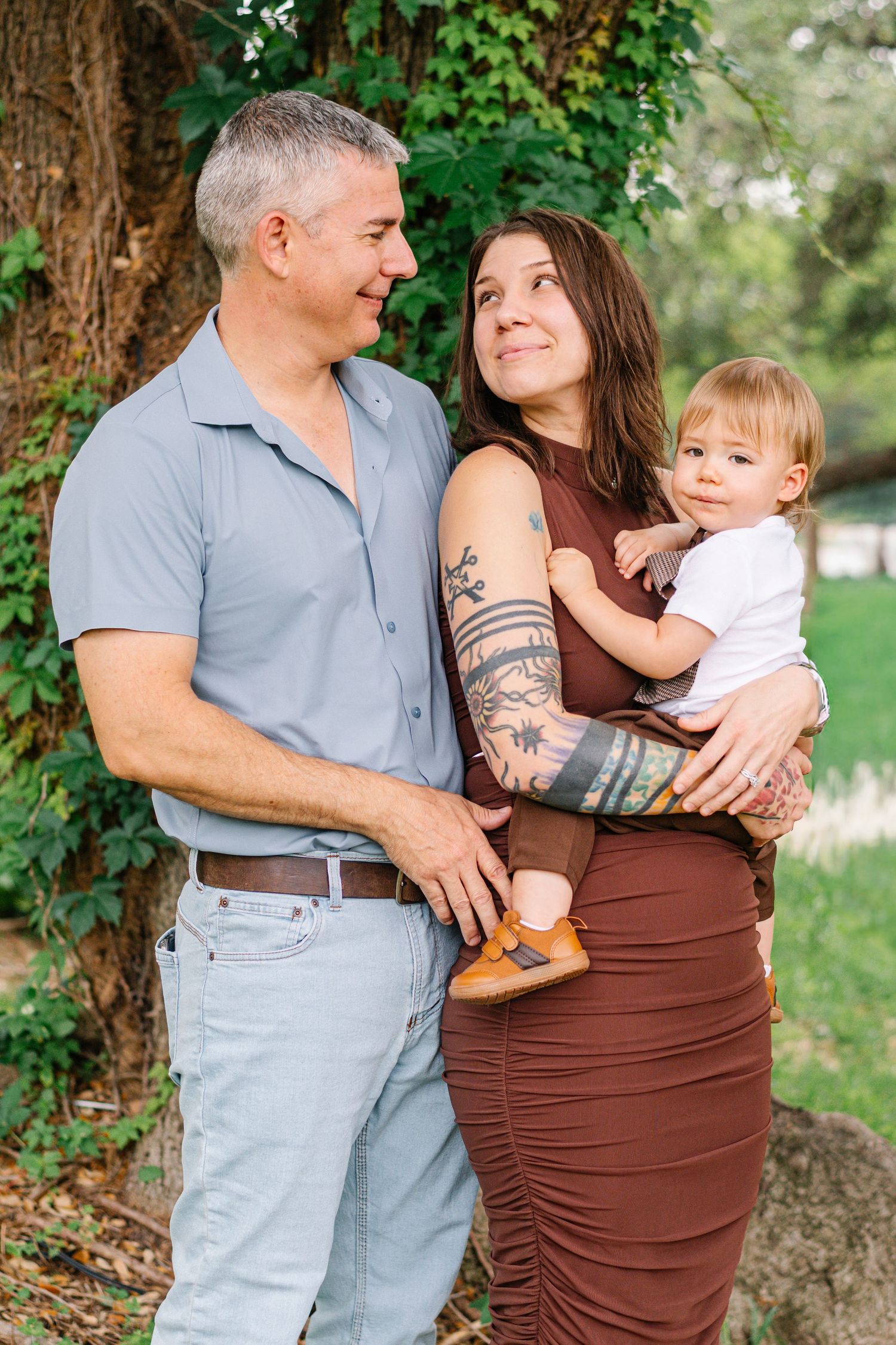 A tattooed woman in a brown dress holds a baby while a gray-haired man smiles at her outdoors.