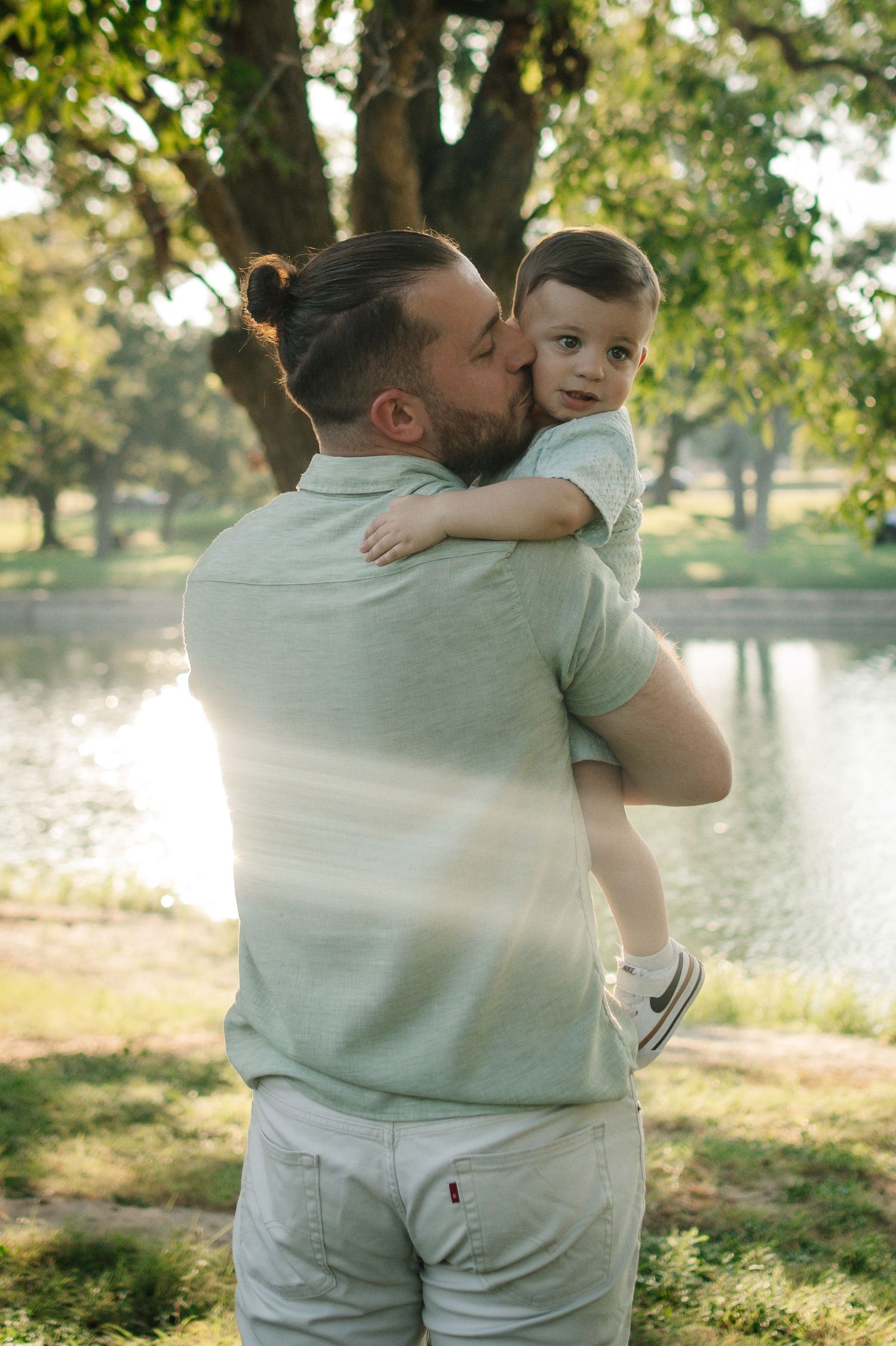 A bearded father kisses his young toddler son while holding him in a sunlit park near a pond.