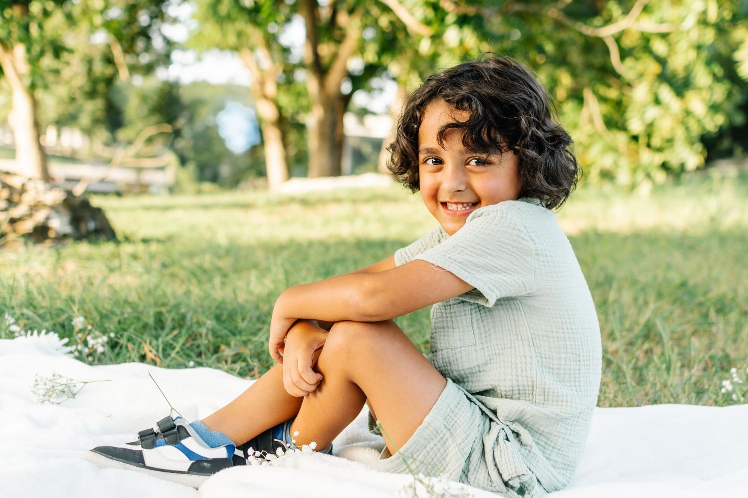 A smiling child with curly hair sits on a blanket in a sunny park, hugging their knees and wearing a light outfit.