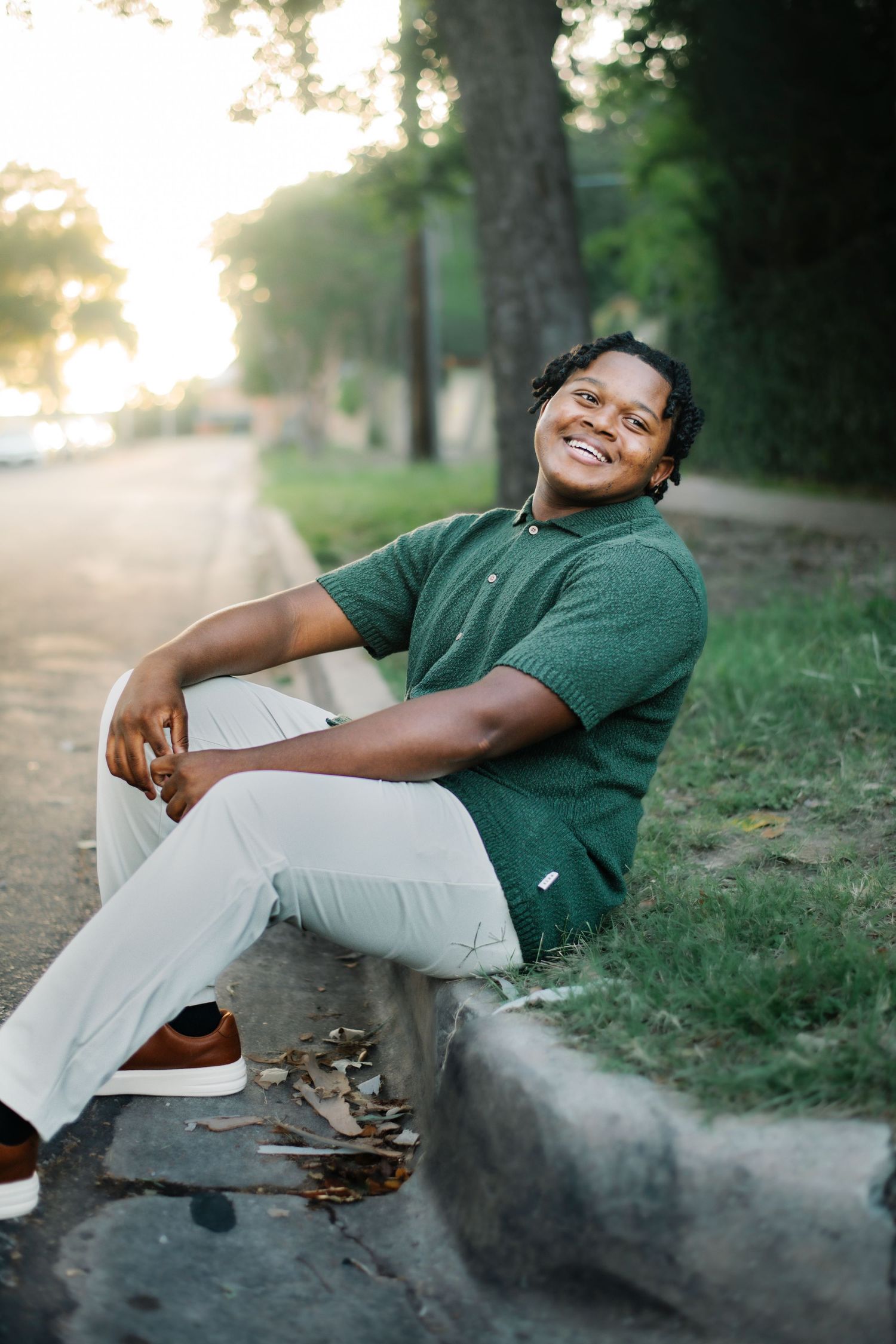 Young man in green polo and white pants smiling while sitting on a curb outdoors at golden hour.