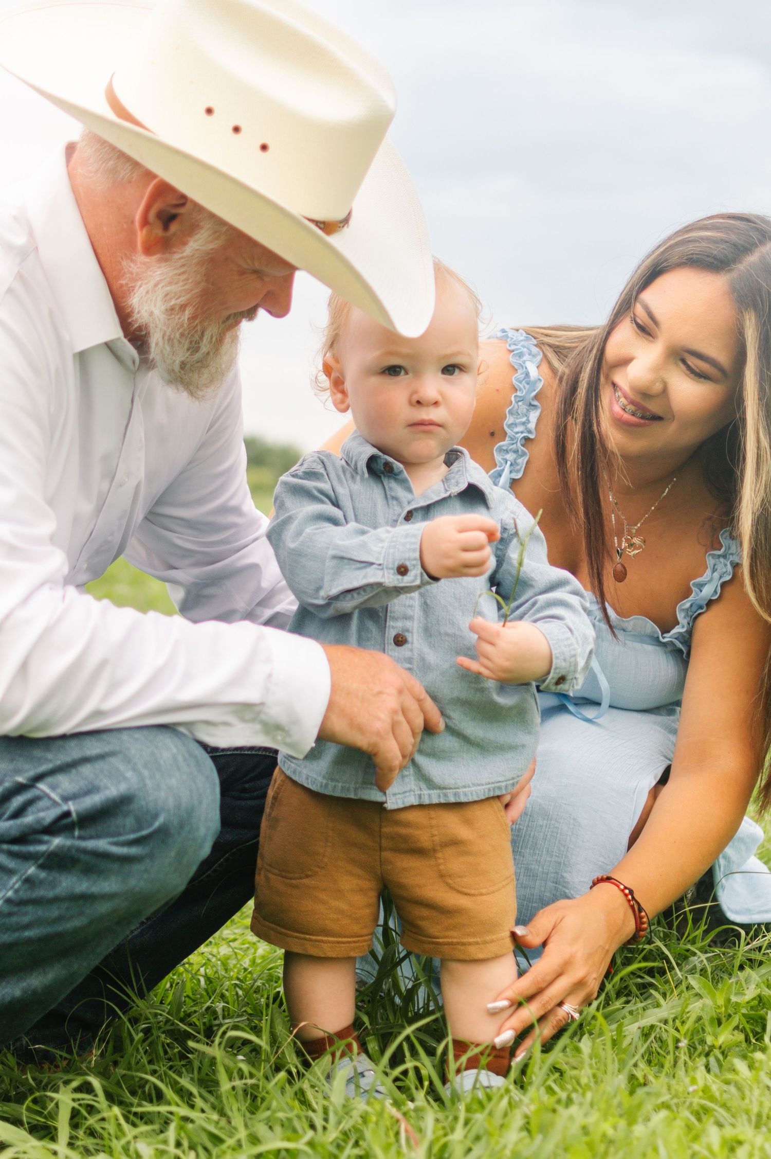 Elderly man in cowboy hat and young woman helping a baby take first steps on green grass outdoors.