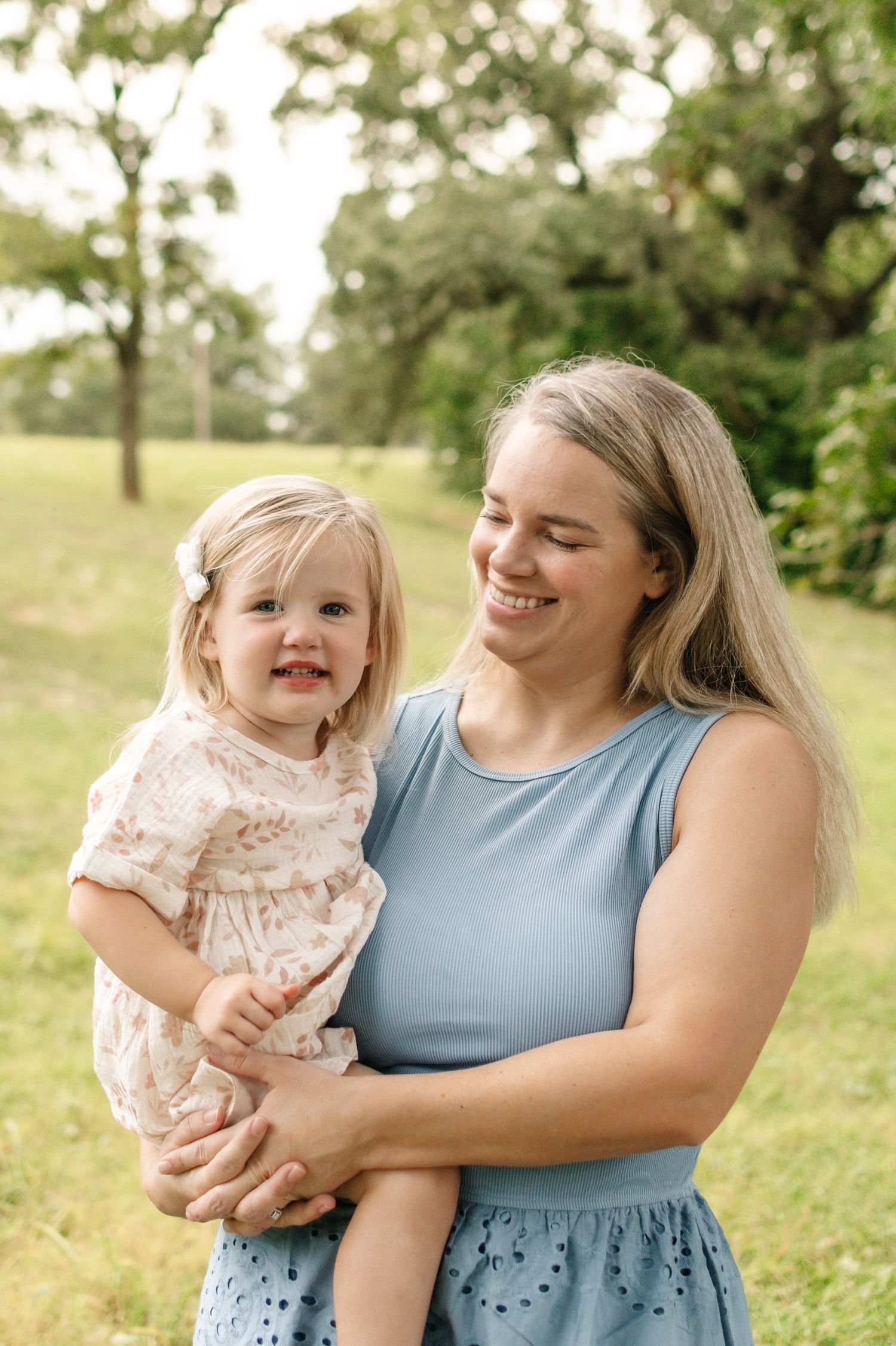 A smiling woman in a blue dress holds a happy toddler girl in a floral dress outdoors in a green park.