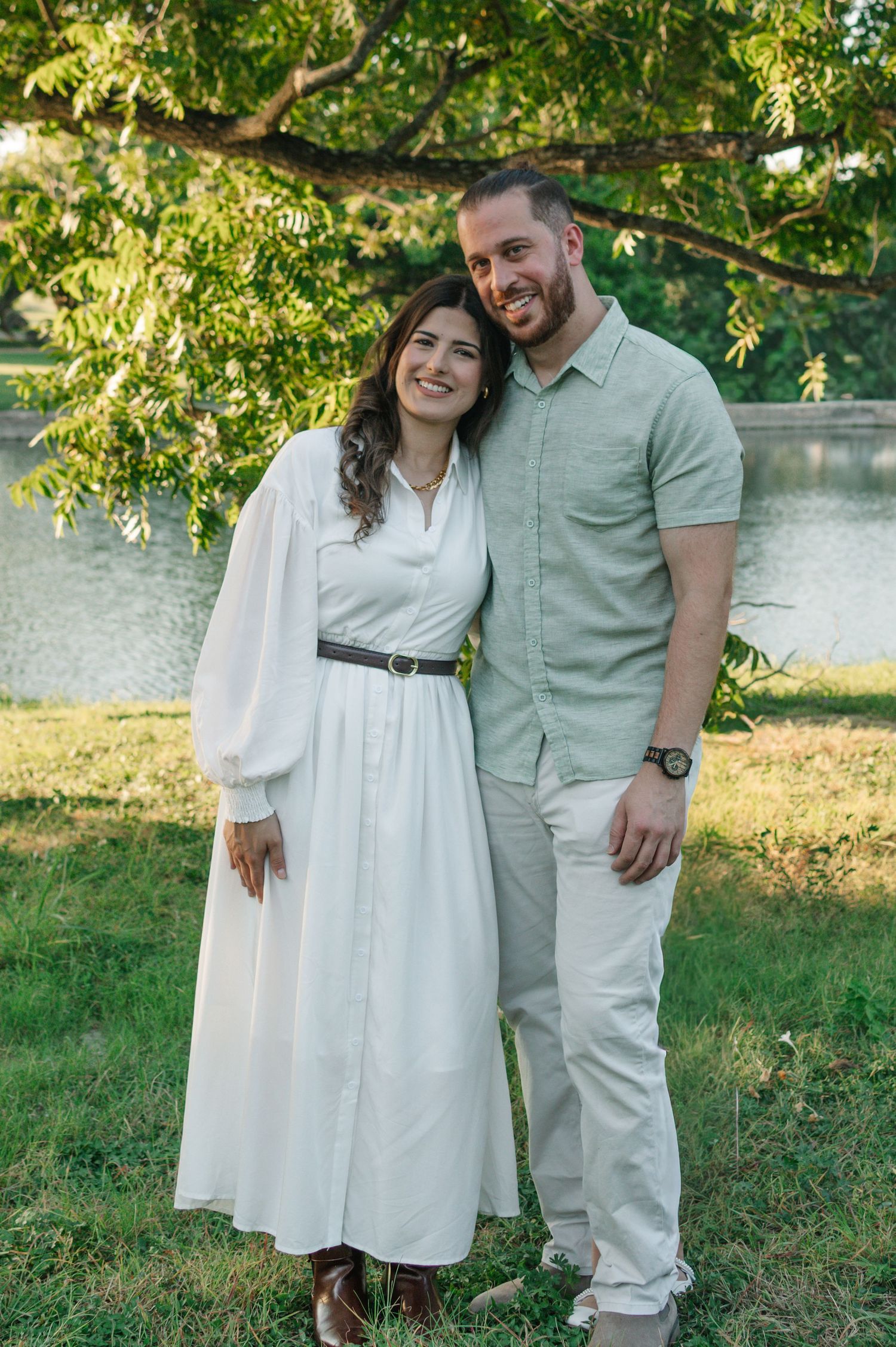 Smiling couple posing outdoors near a pond, woman in white dress with belt, man in light green shirt and khakis.