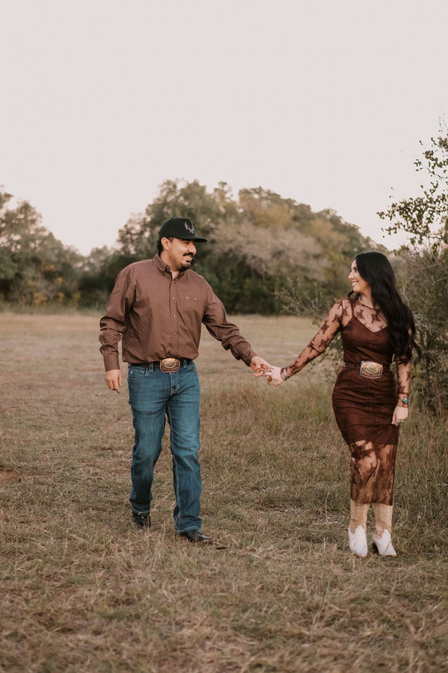 A couple holds hands walking through a rustic field, dressed in coordinating brown western outfits with boots and a cap.