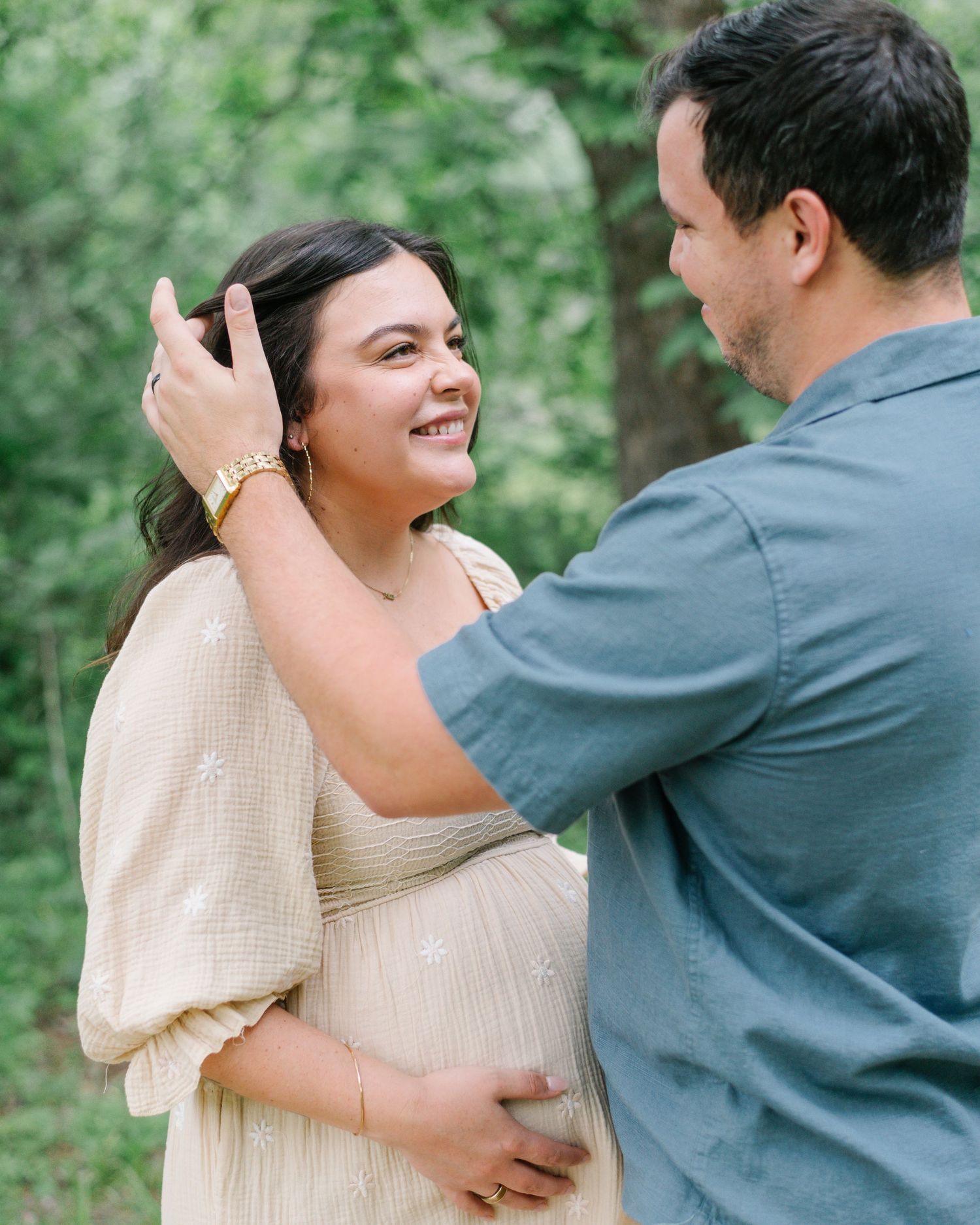 Pregnant woman in cream dress smiles as partner tenderly brushes her hair in a lush green outdoor setting.