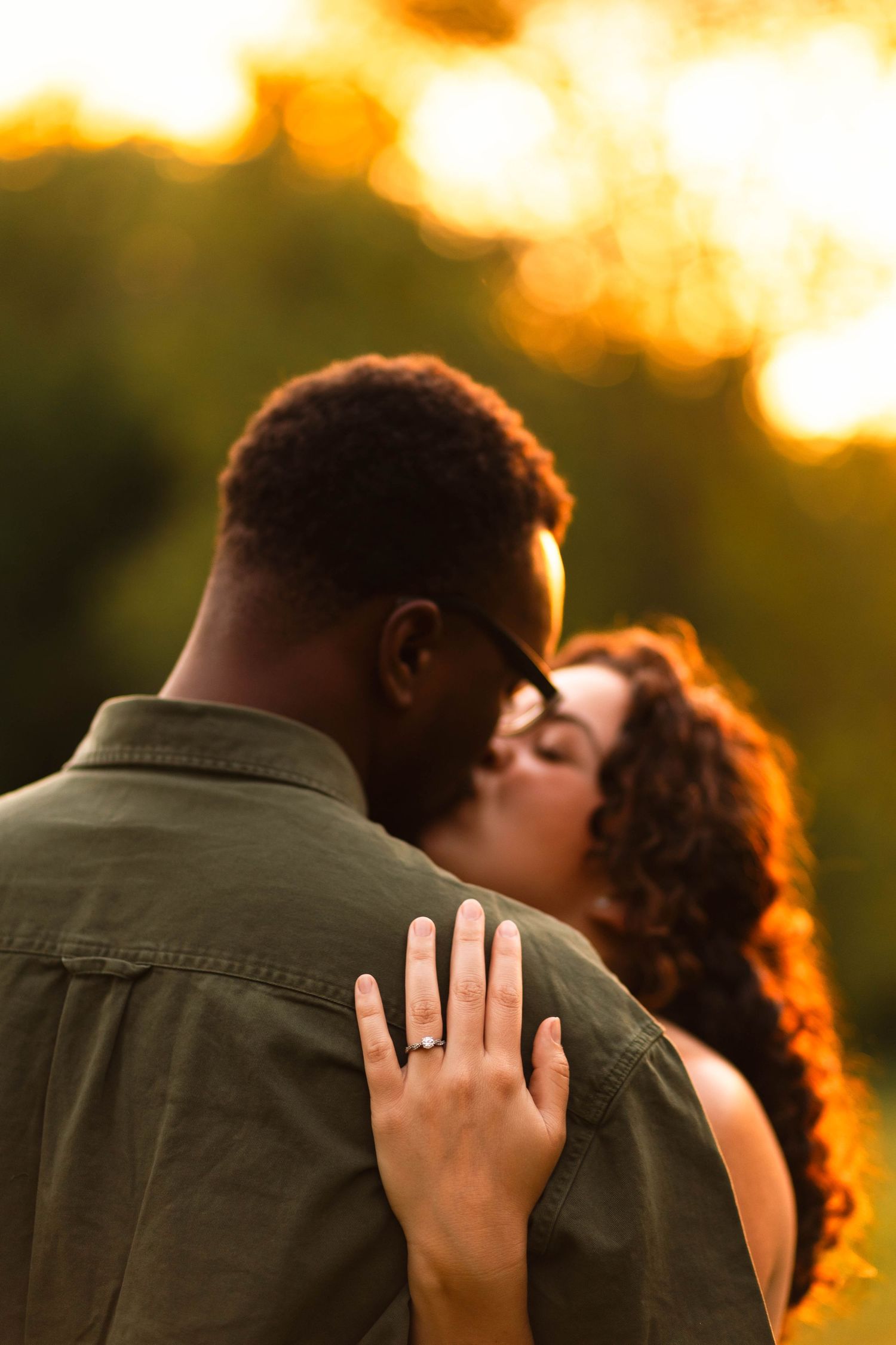 Couple sharing a romantic kiss at golden hour, woman showing engagement ring on man's shoulder at Grings Mill in Reading, PA.