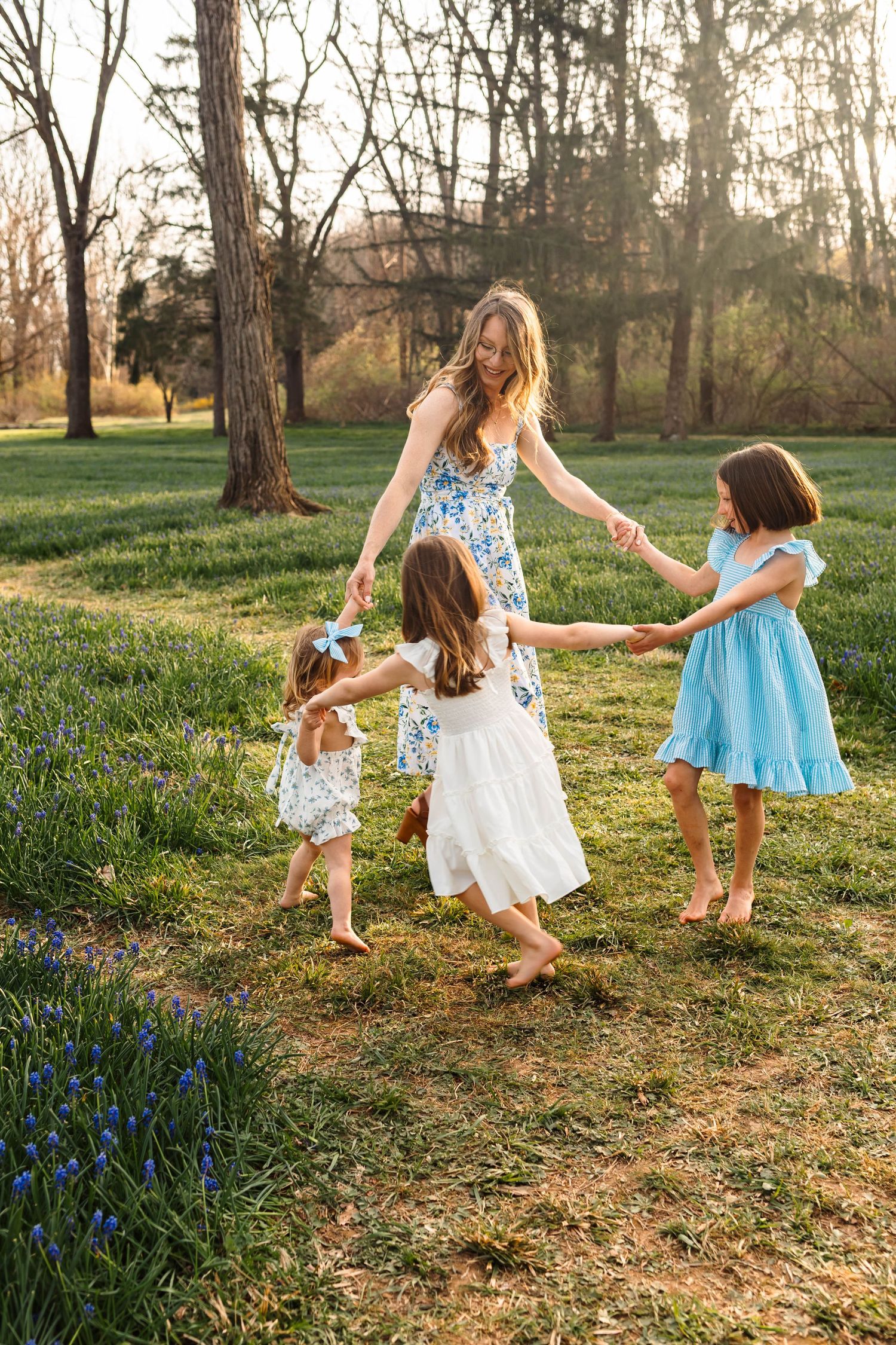 A joyful mother dances in a circle with her three daughters in a sunlit park filled with grape hyacinths in Alburtis, PA at Lockridge Park.