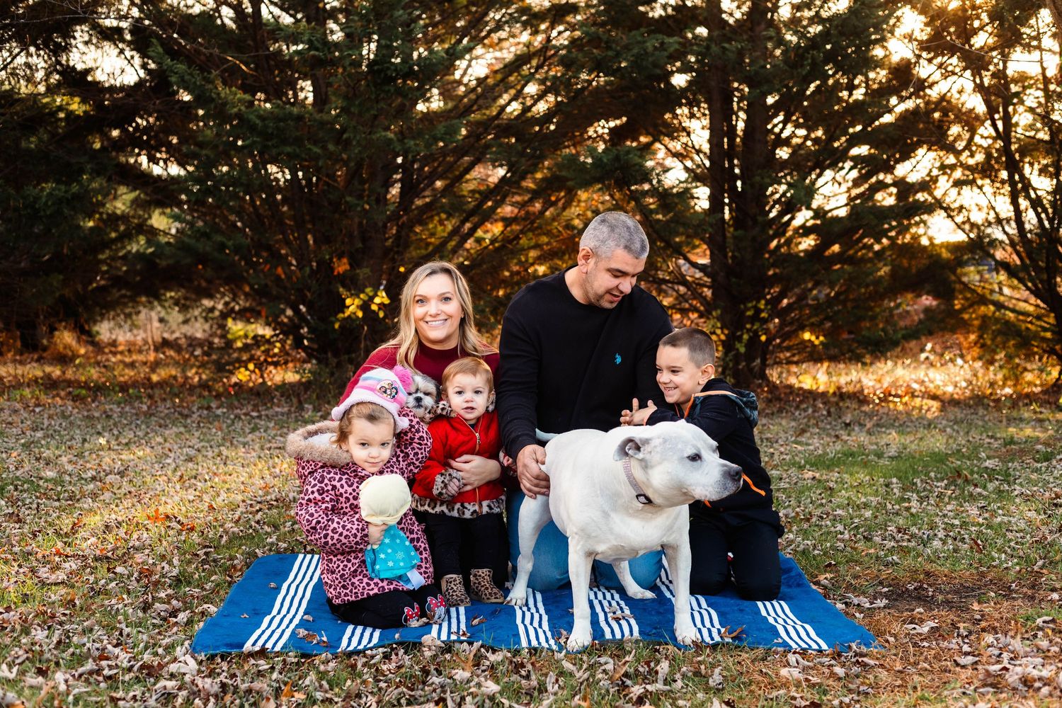 Family of five with white dog sits on blue blanket among autumn trees during golden hour in Boyertown, PA.