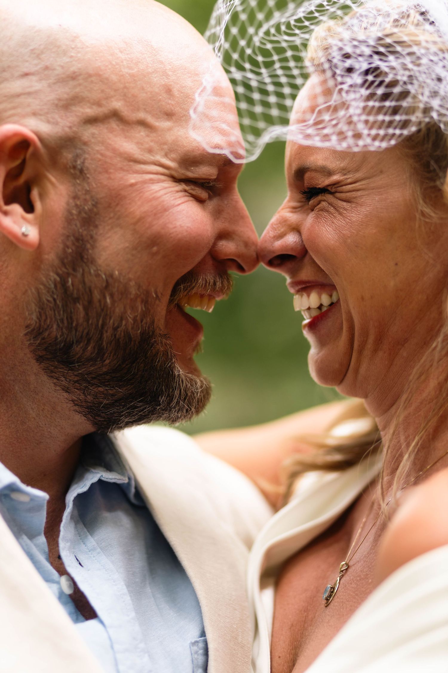 Laughing bride and groom touching noses on wedding day, sharing a joyful intimate moment outdoors.