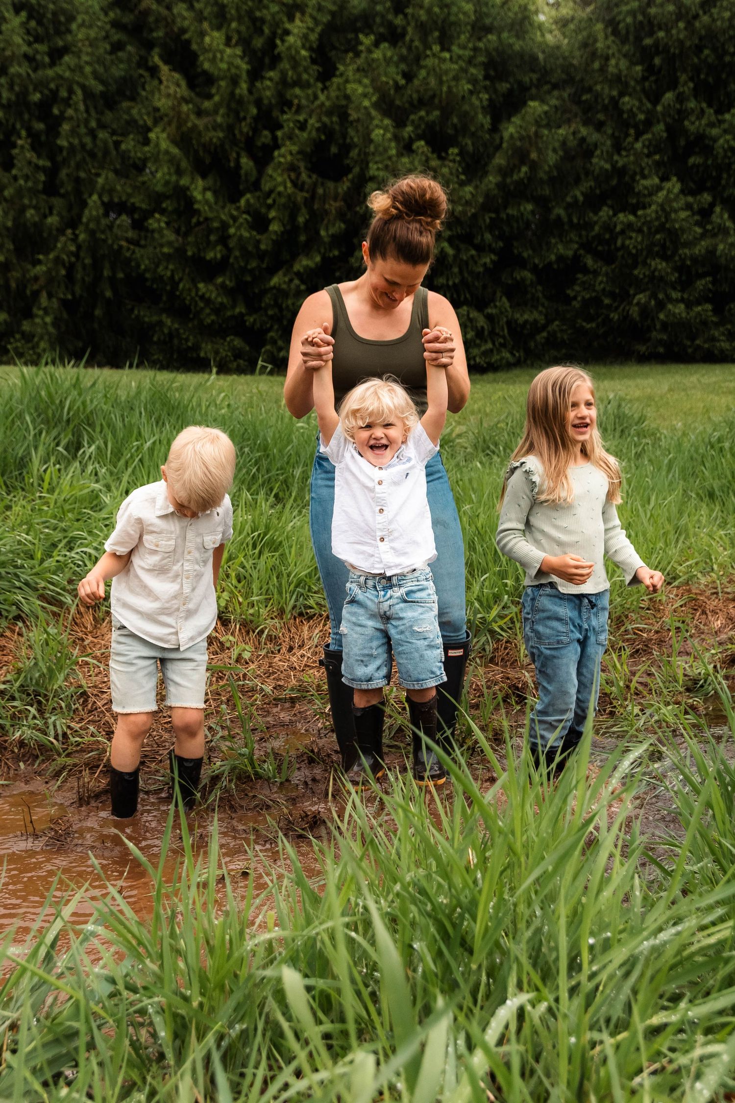 Mother lifts toddler while two other children play in muddy stream surrounded by tall green grass.