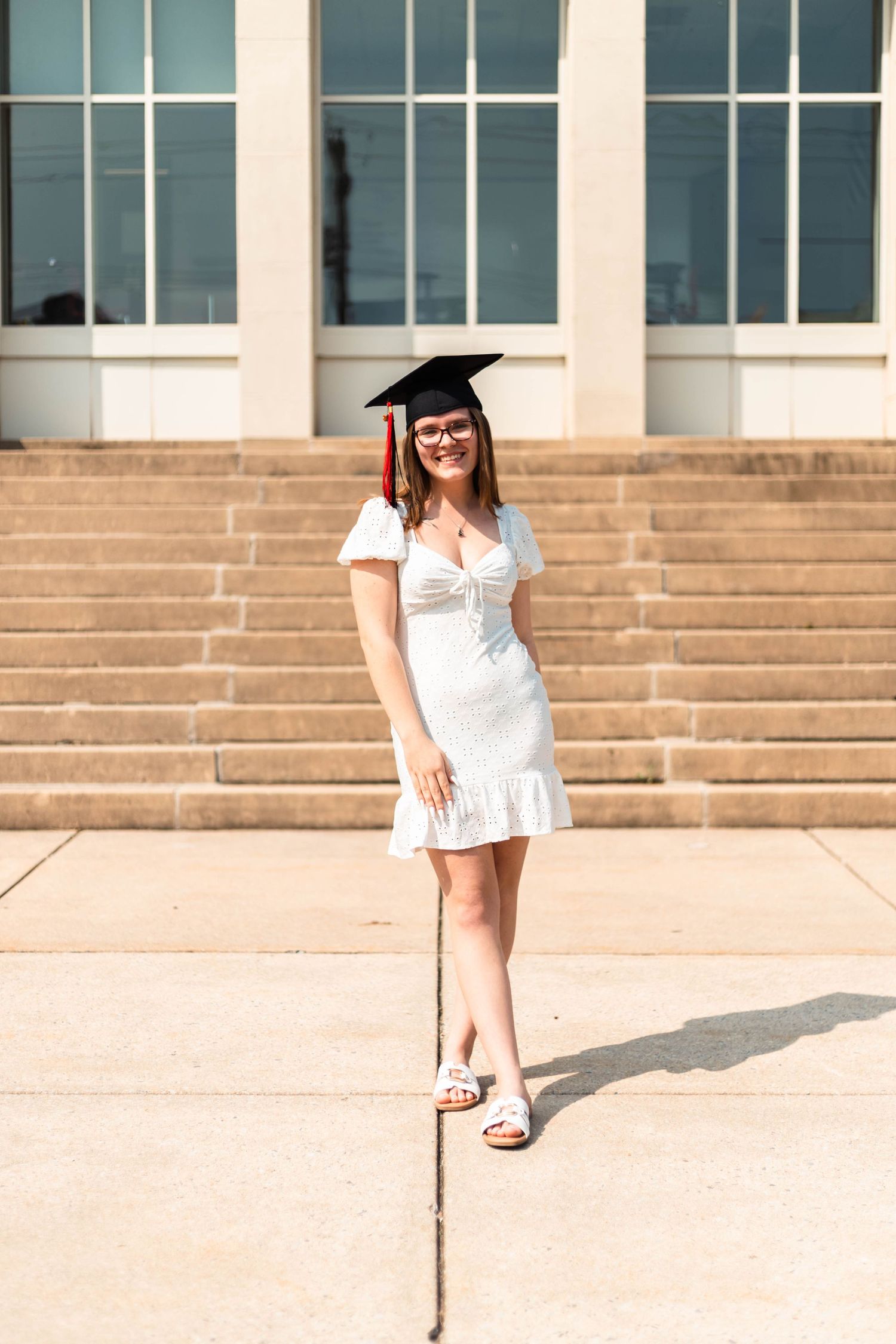 Smiling graduate in white dress and black mortarboard cap stands on steps outside a university building.