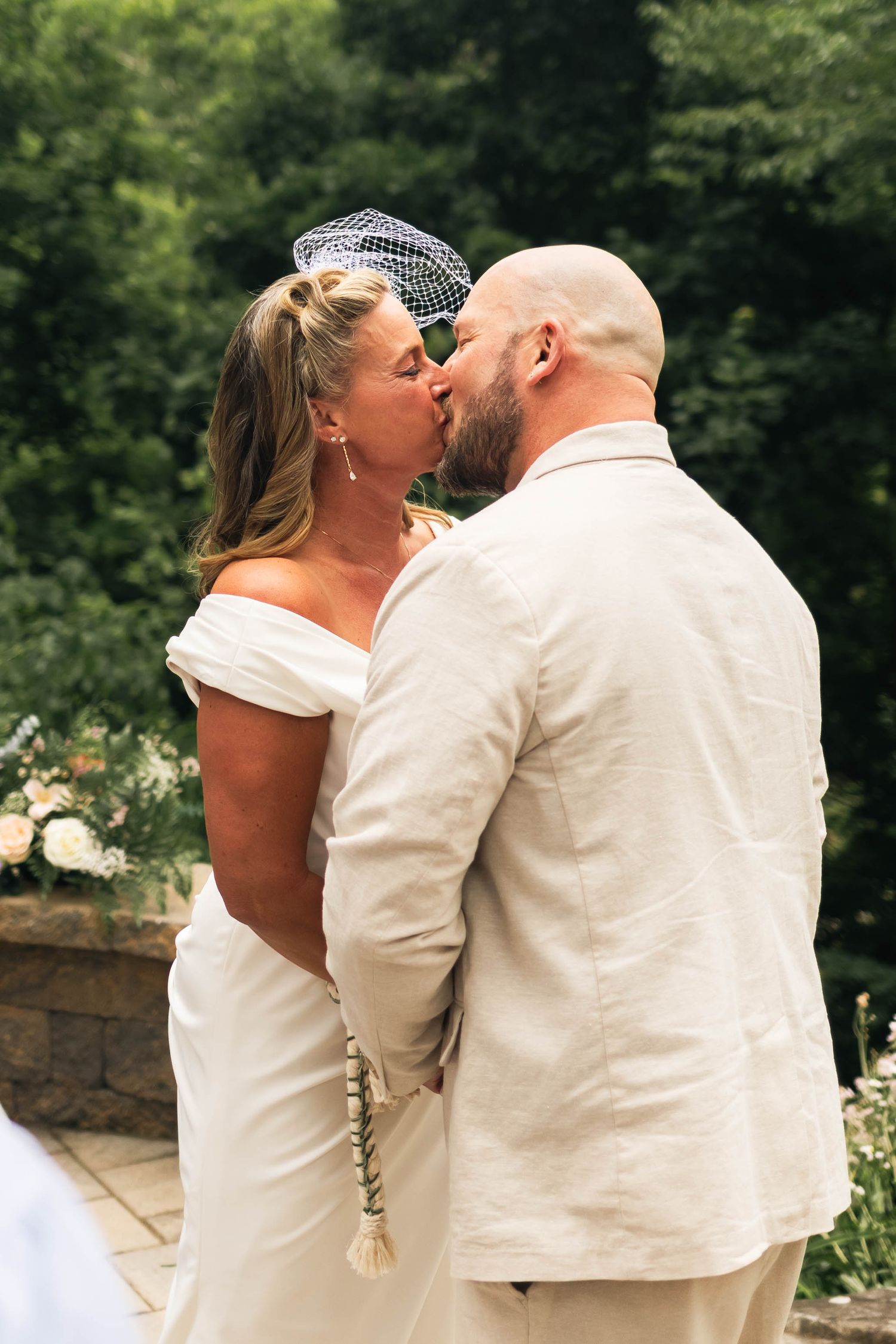 Bride and bald groom share first kiss at outdoor wedding ceremony surrounded by lush green trees.