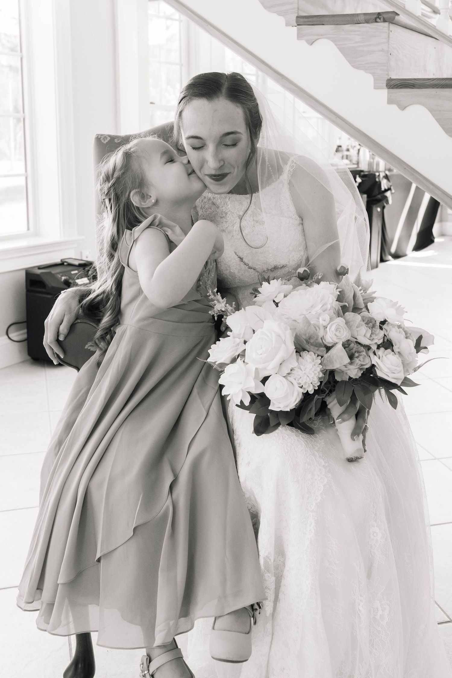 Sweet flower girl in grey dress gives bride a kiss on the cheek in black and white photo.