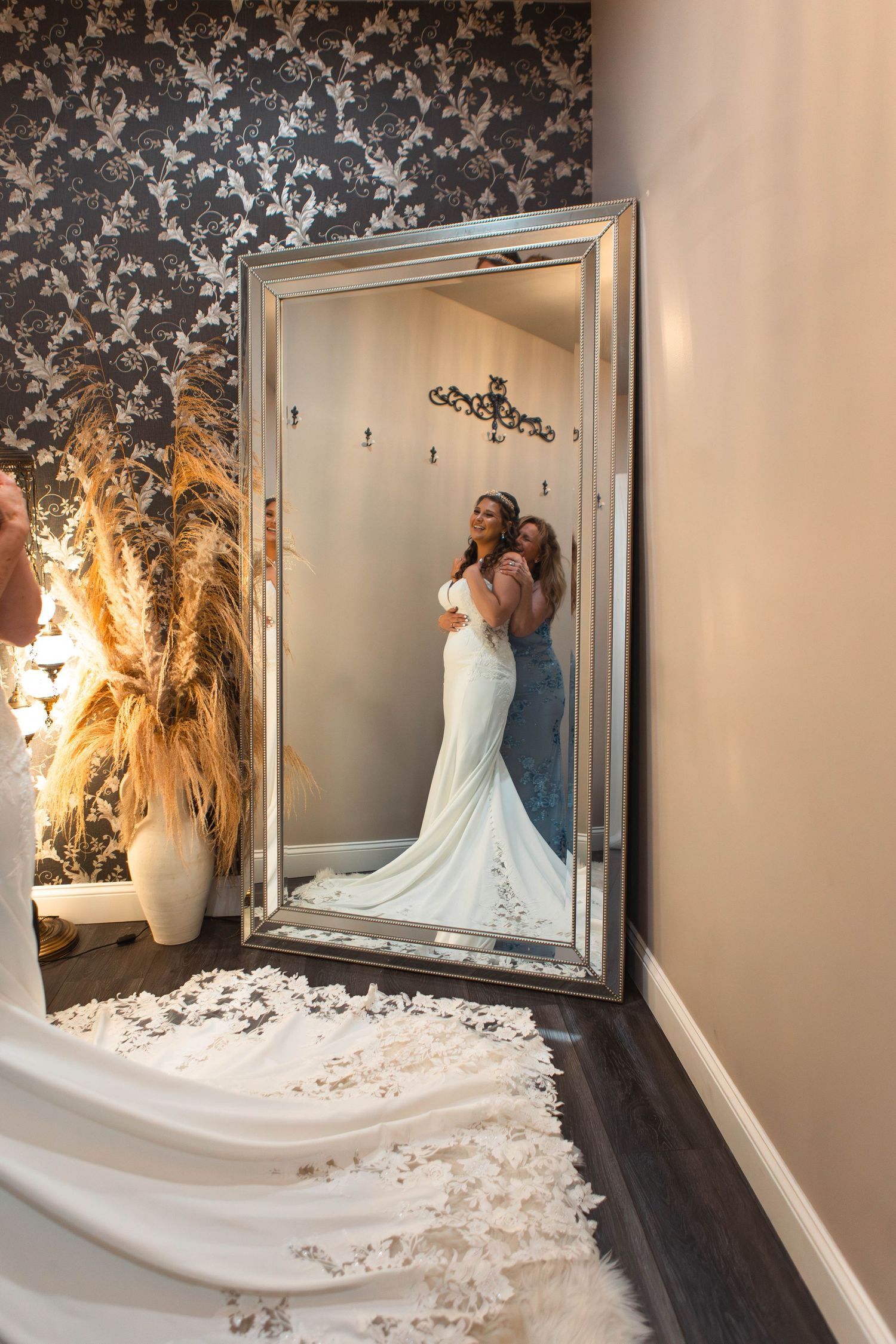 Bride in white mermaid gown admires her reflection in an ornate floor-length mirror in a bridal boutique.