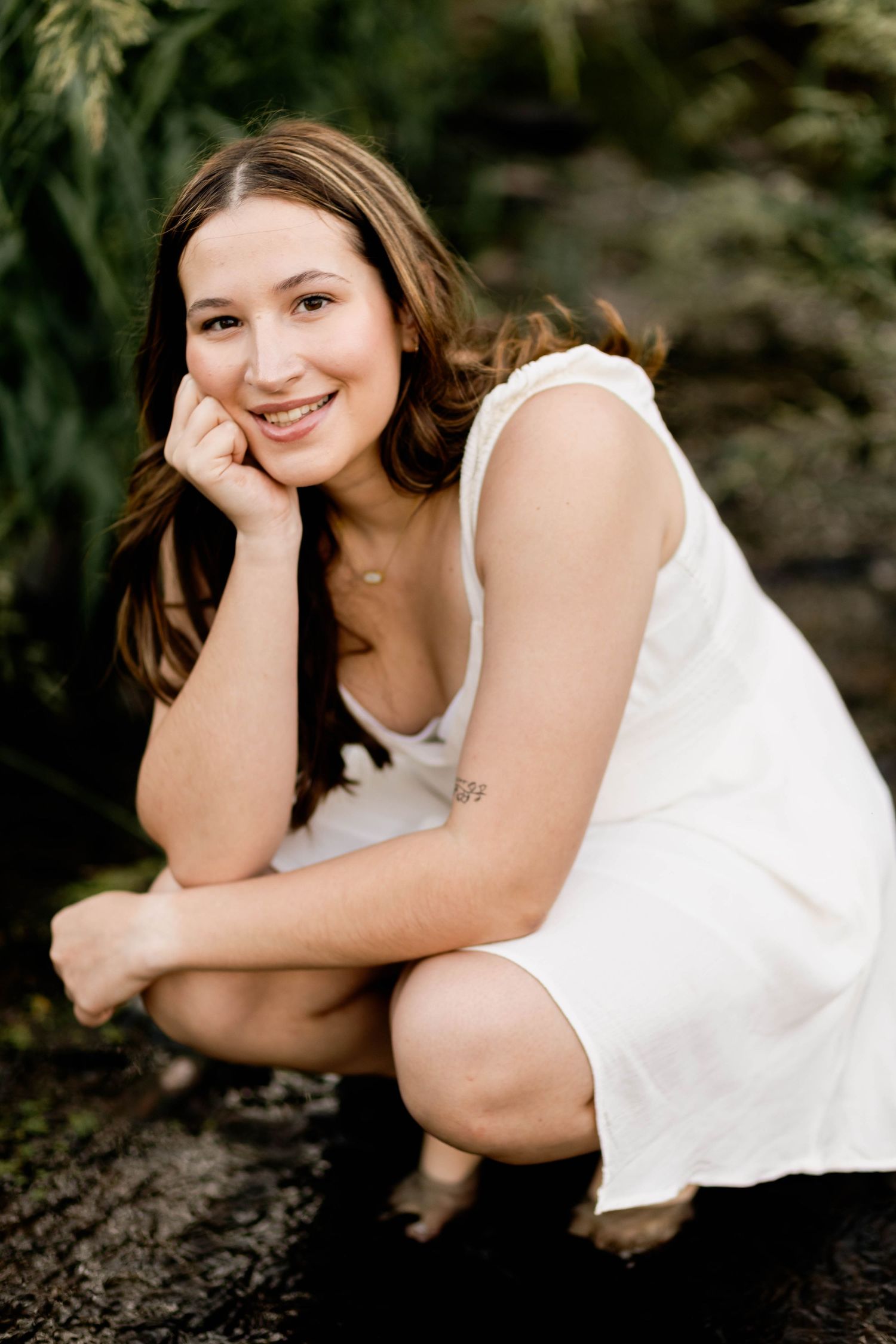 Boyertown High School Senior in white dress crouches near greenery in senior portrait.