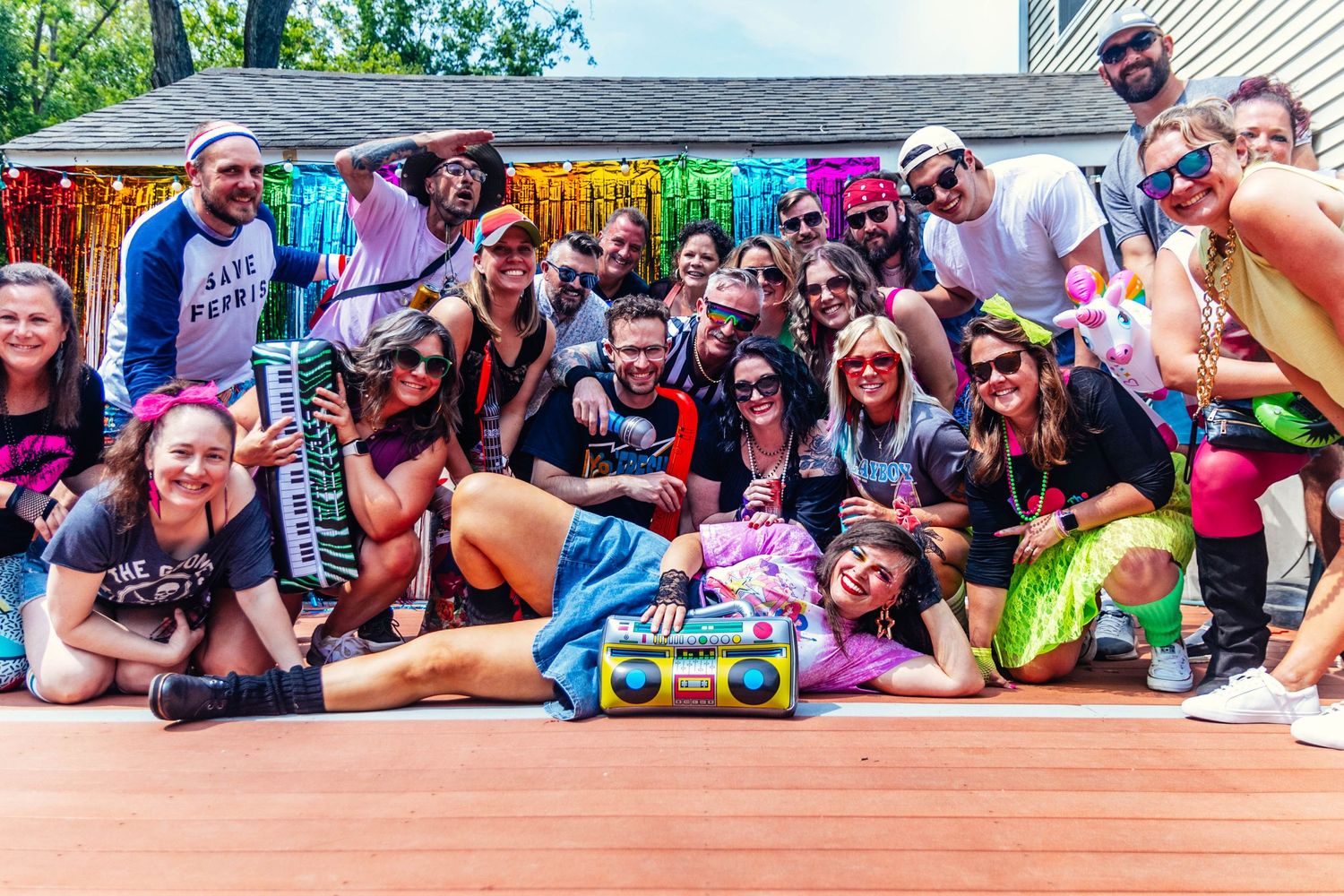 Large group of friends posing together at a colorful 80s-themed outdoor birthday party celebration.